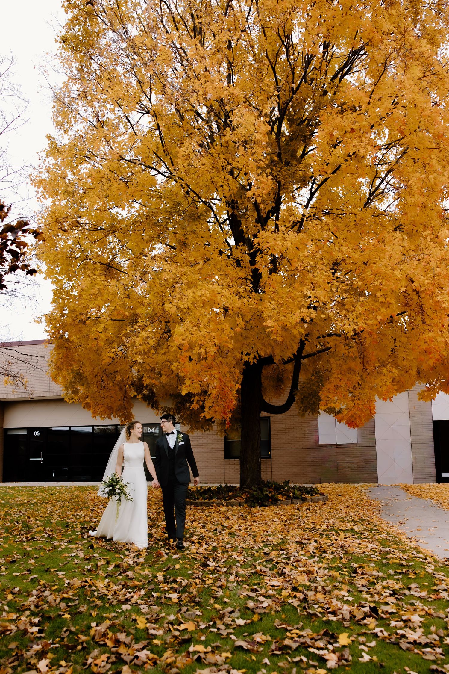 Wedding couple stands beneath vibrant golden autumn tree with fallen leaves.