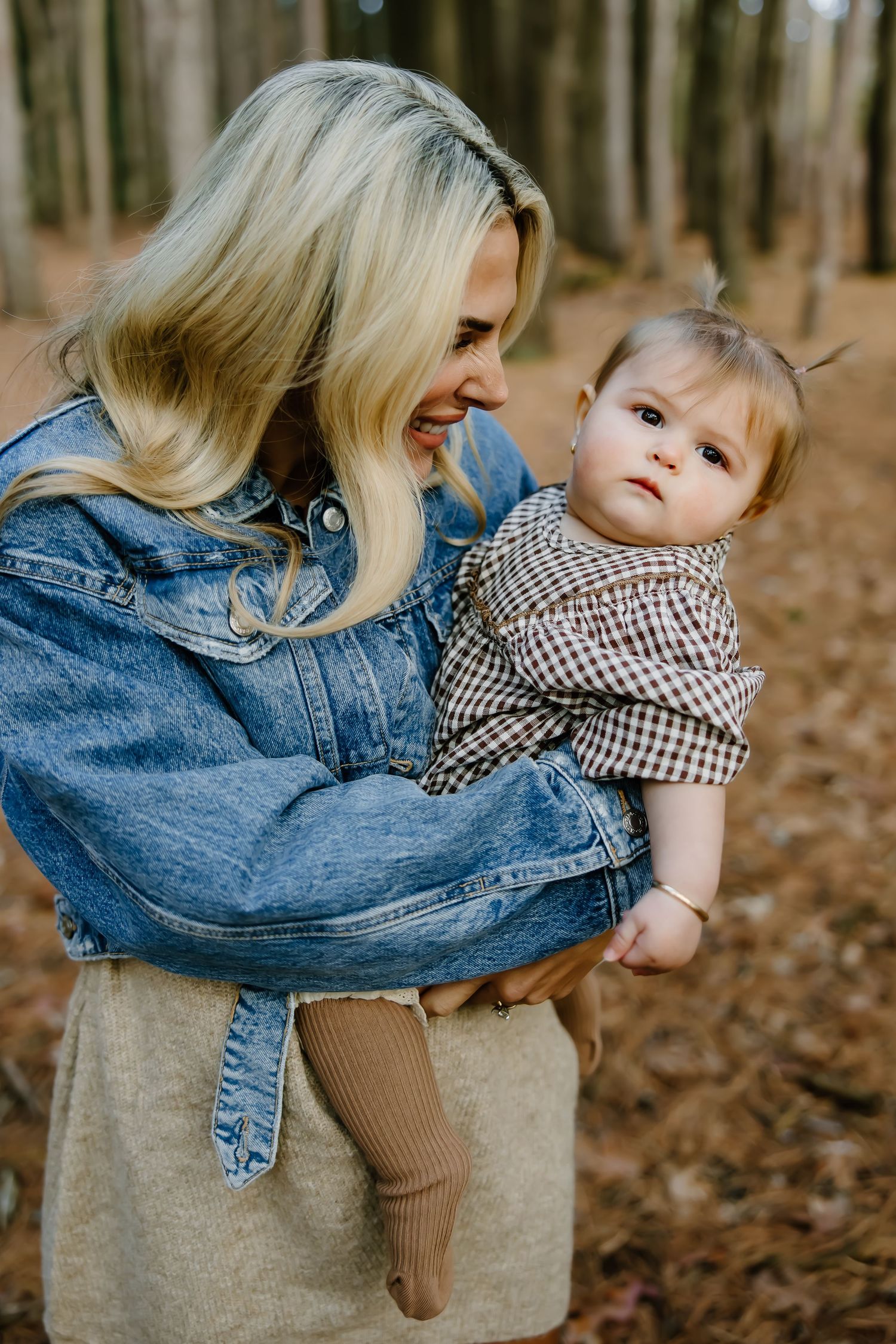 Parent in denim jacket holding young child in checkered outfit among trees.