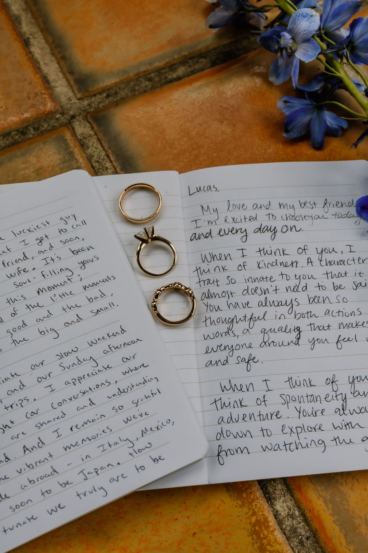 Three gold wedding rings rest on handwritten vow letters beside blue flowers on terracotta tiles.