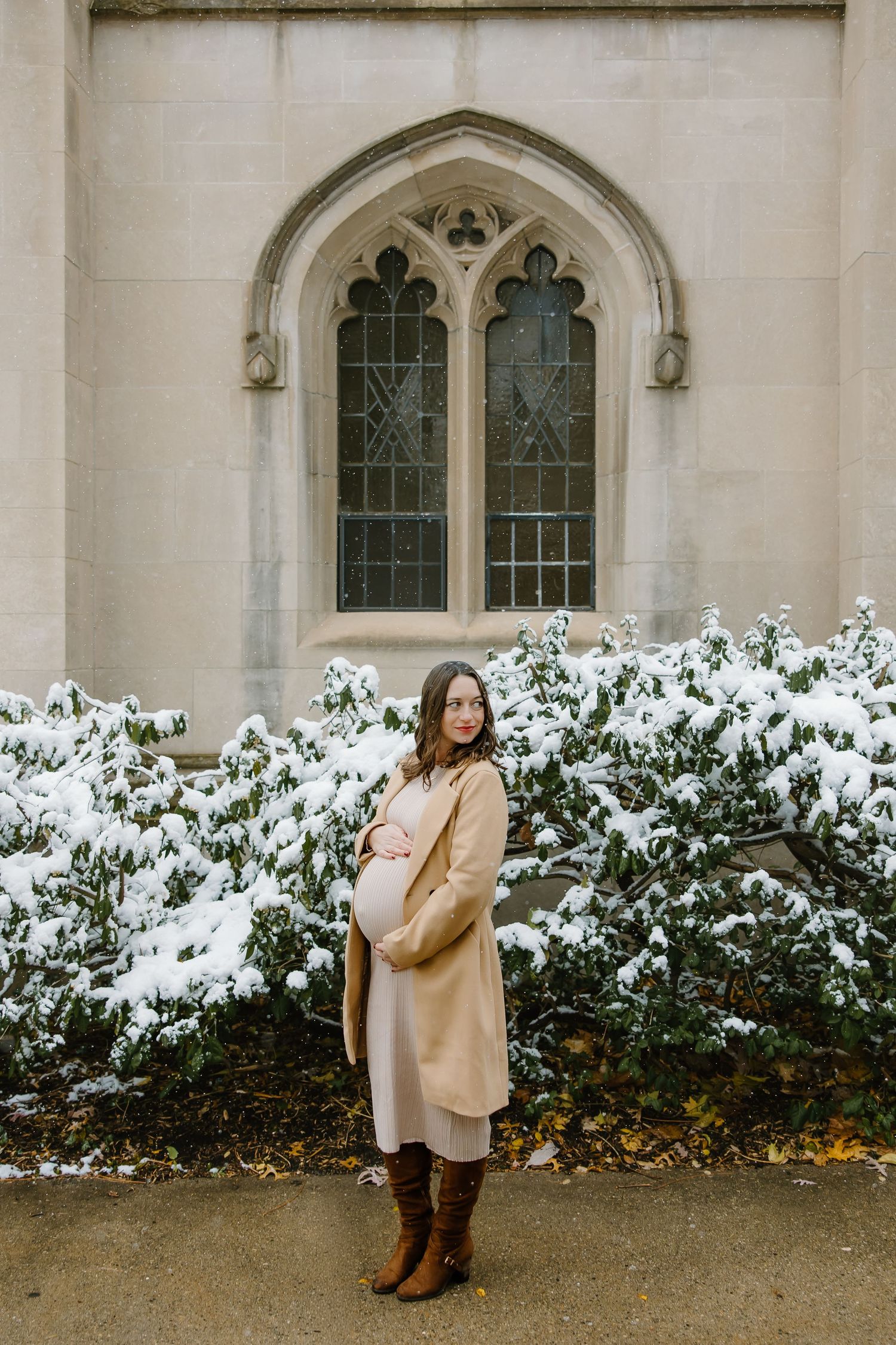 A pregnant woman in a beige coat poses in front of a snow-covered Gothic church window.