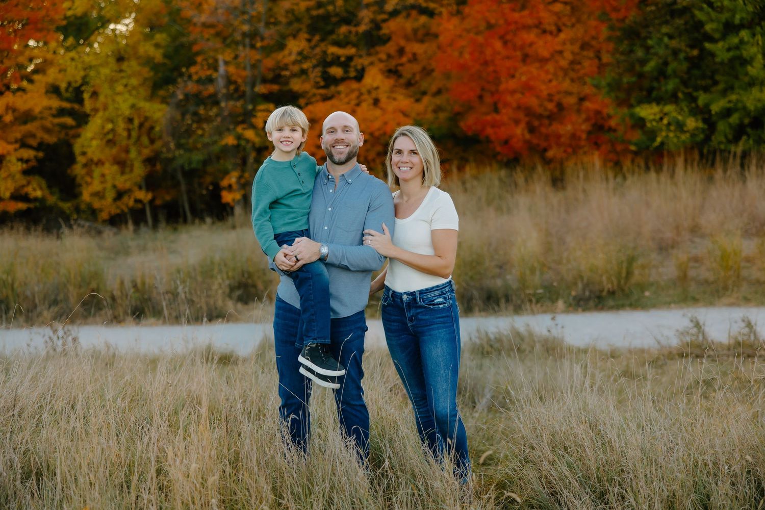 Couple embraces in a field surrounded by vibrant autumn foliage and golden grass at sunset.