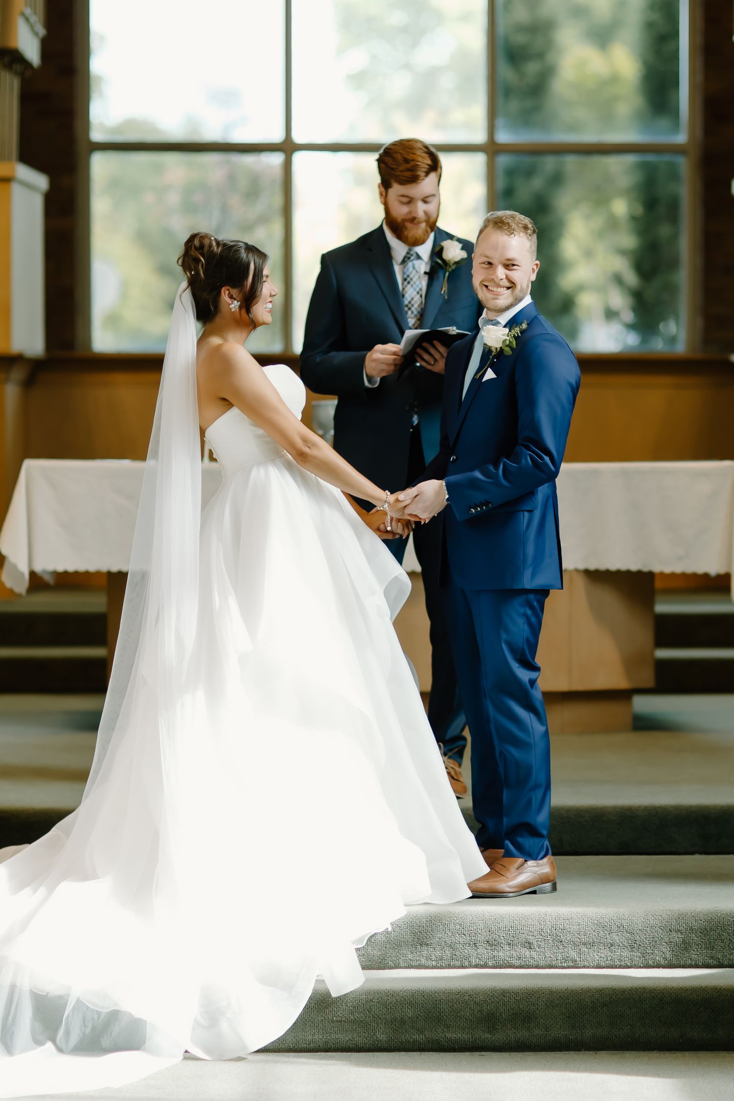 A wedding ceremony takes place on steps inside a bright sunlit church sanctuary.