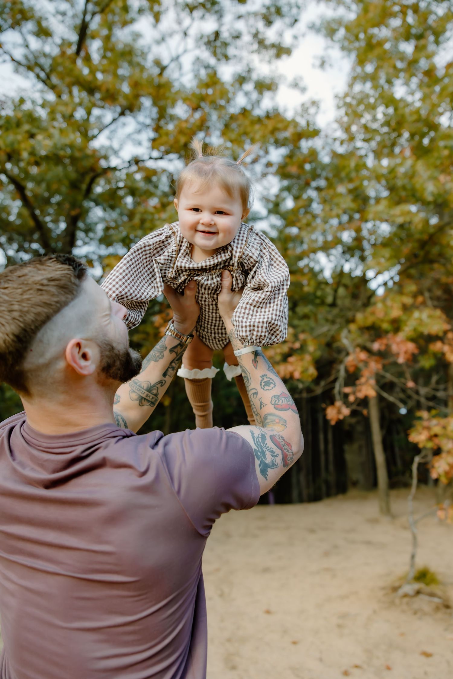 Tattooed person in purple shirt lifts smiling toddler in gingham dress outdoors.