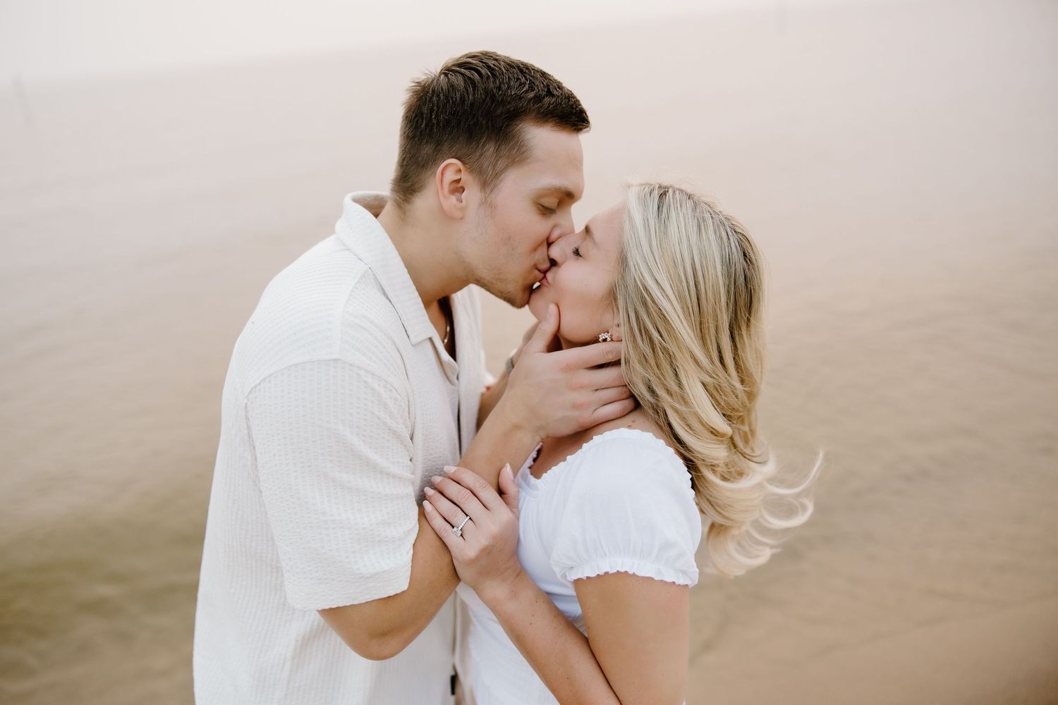 Romantic beach engagement photo with couple in white and khaki embracing.