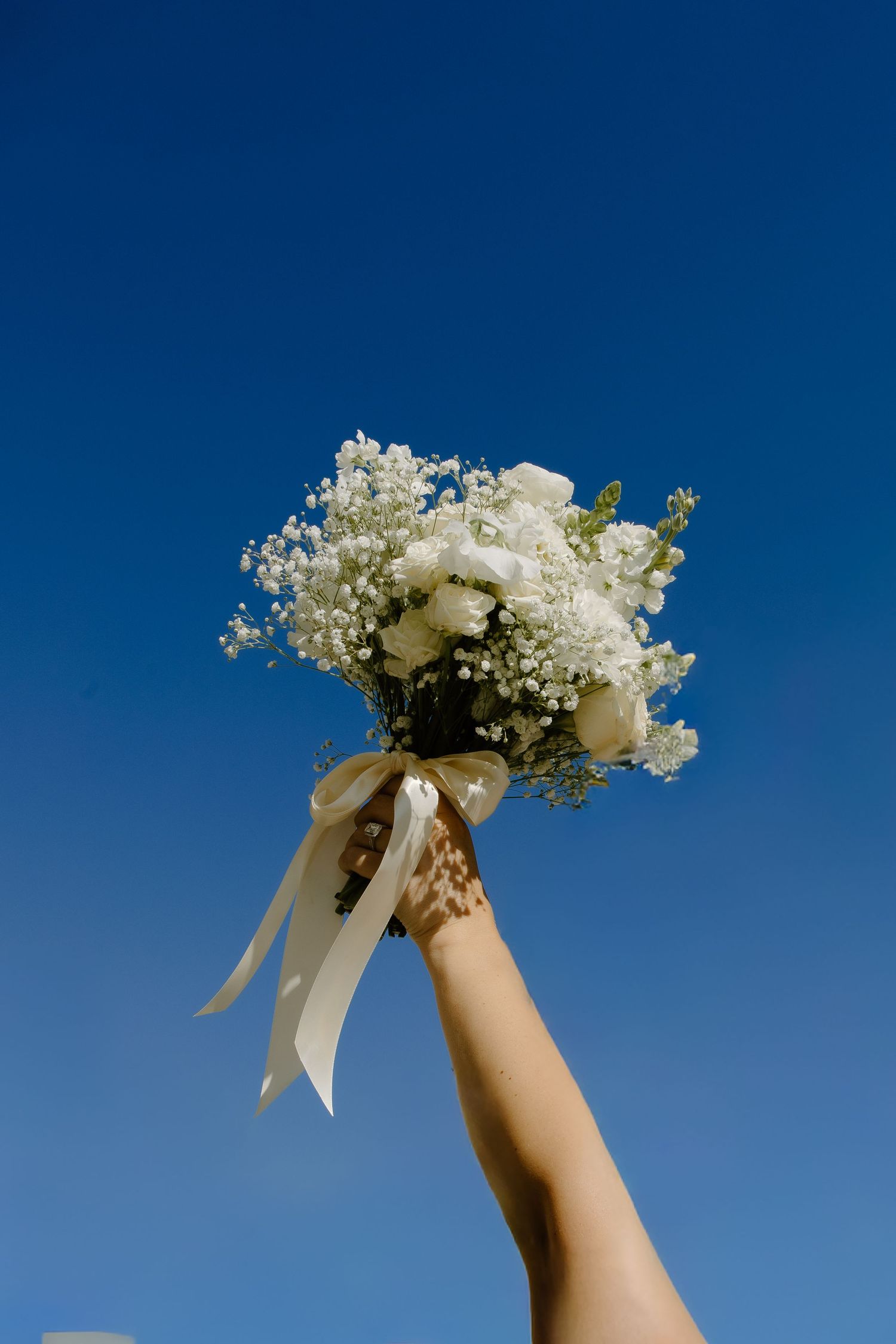 White bridal bouquet with baby's breath flowers held up against bright blue sky with cream colored ribbon.