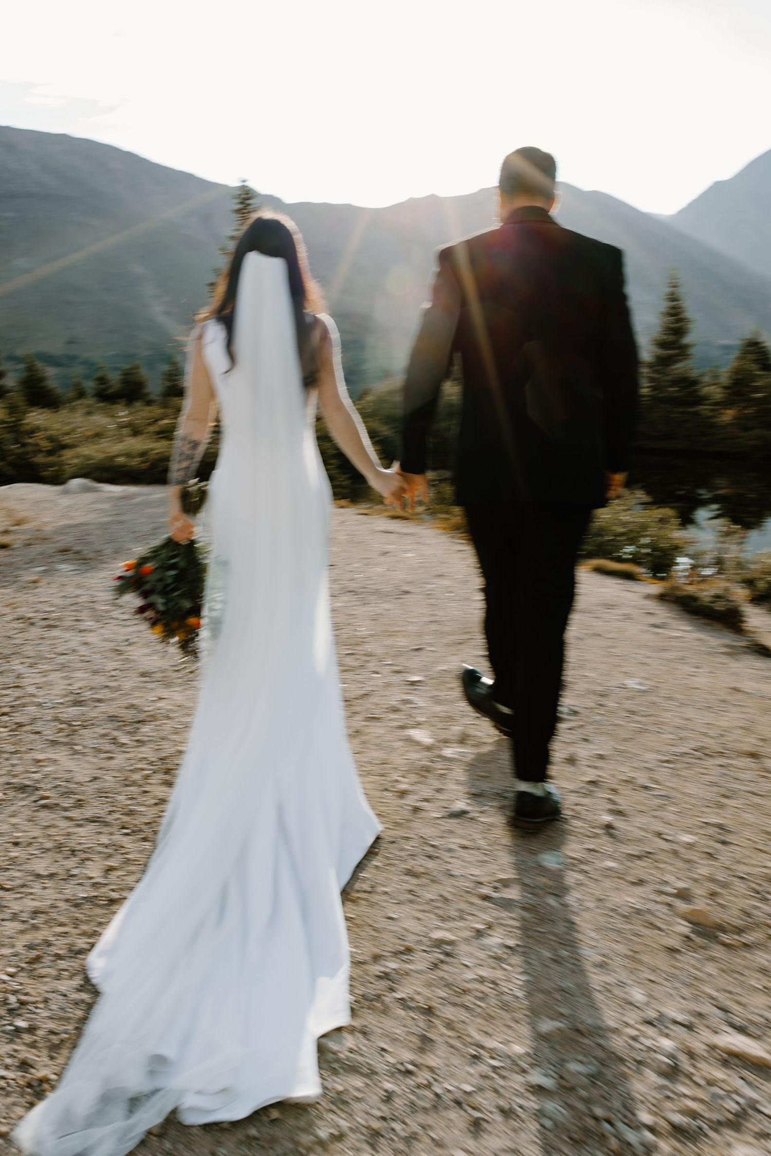 A bride and groom walk hand in hand along a mountain trail at sunset.