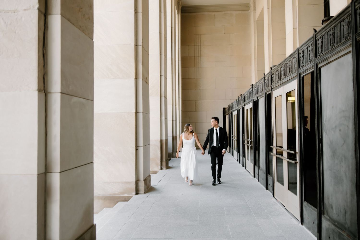 A bride and groom walk hand in hand through an elegant columned corridor in their wedding attire.
