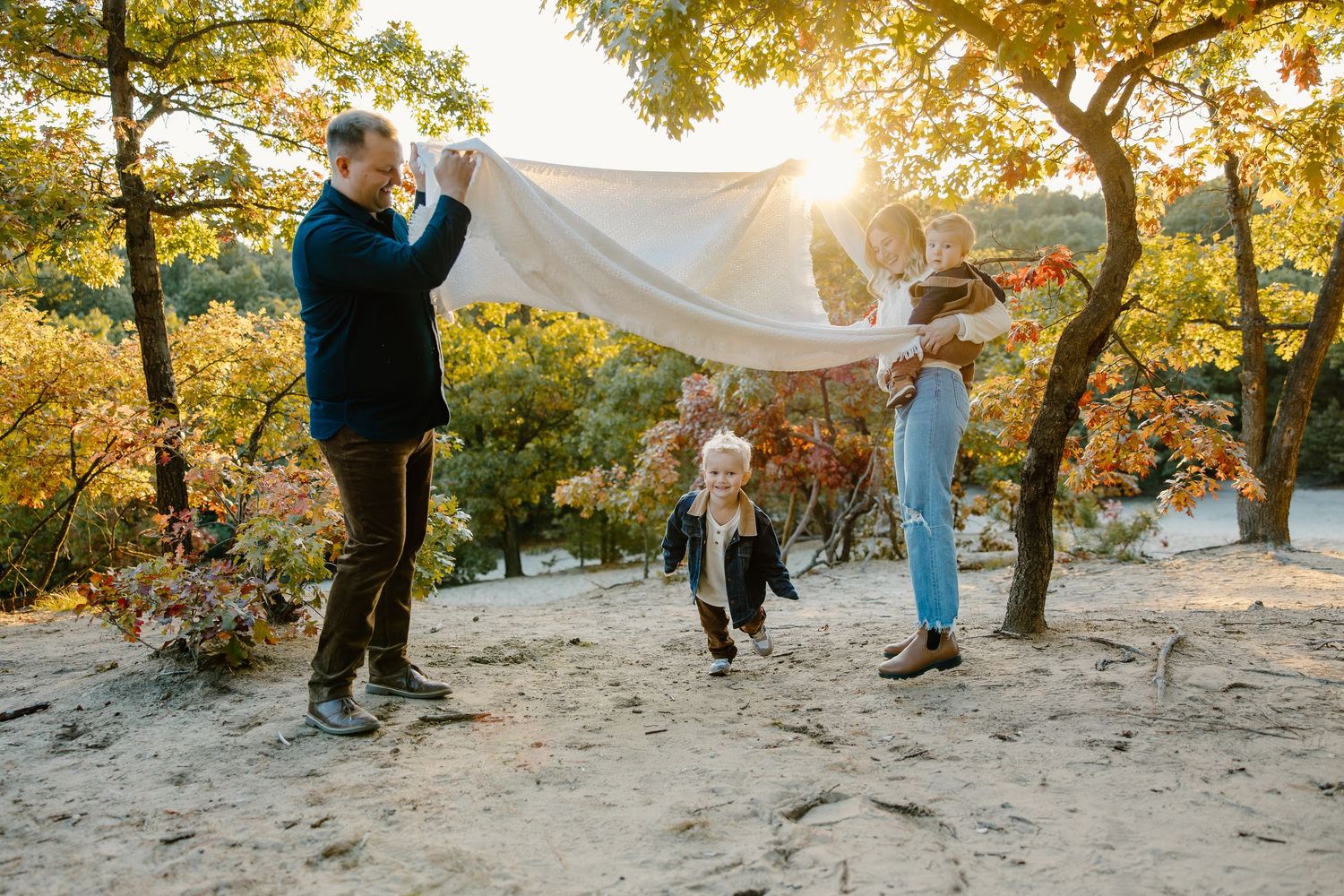Family plays together on sandy ground surrounded by autumn trees at sunset.