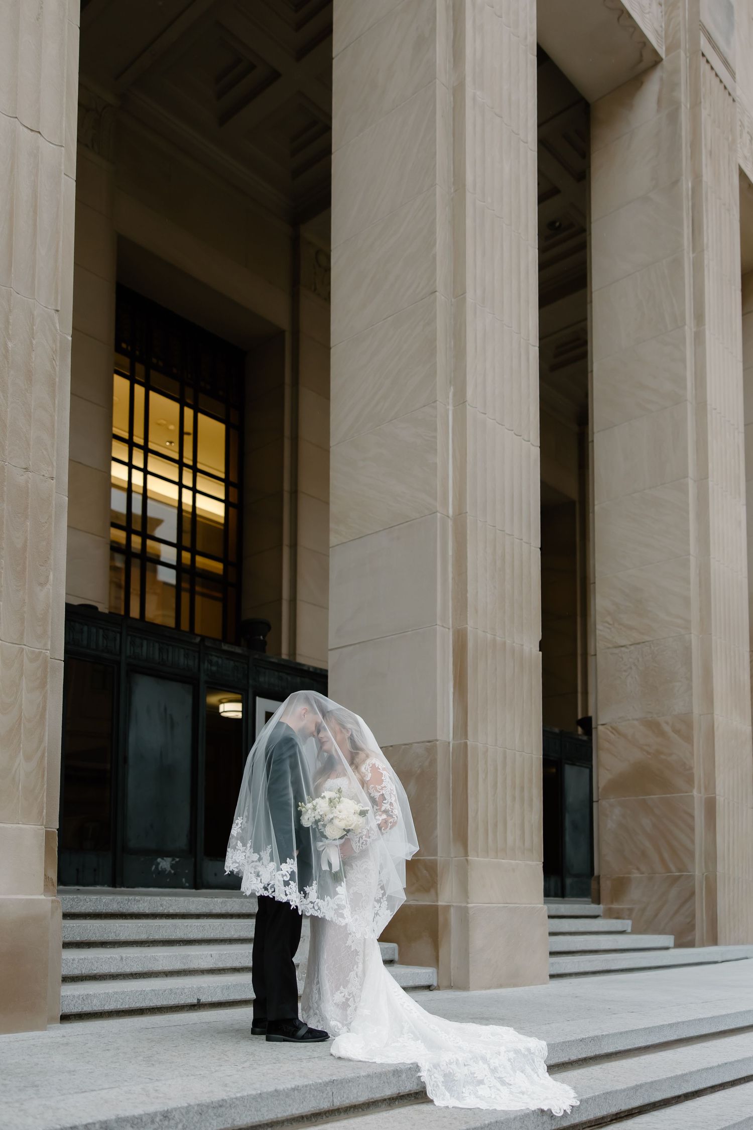 White wedding dress train cascades down marble steps between classical columns.