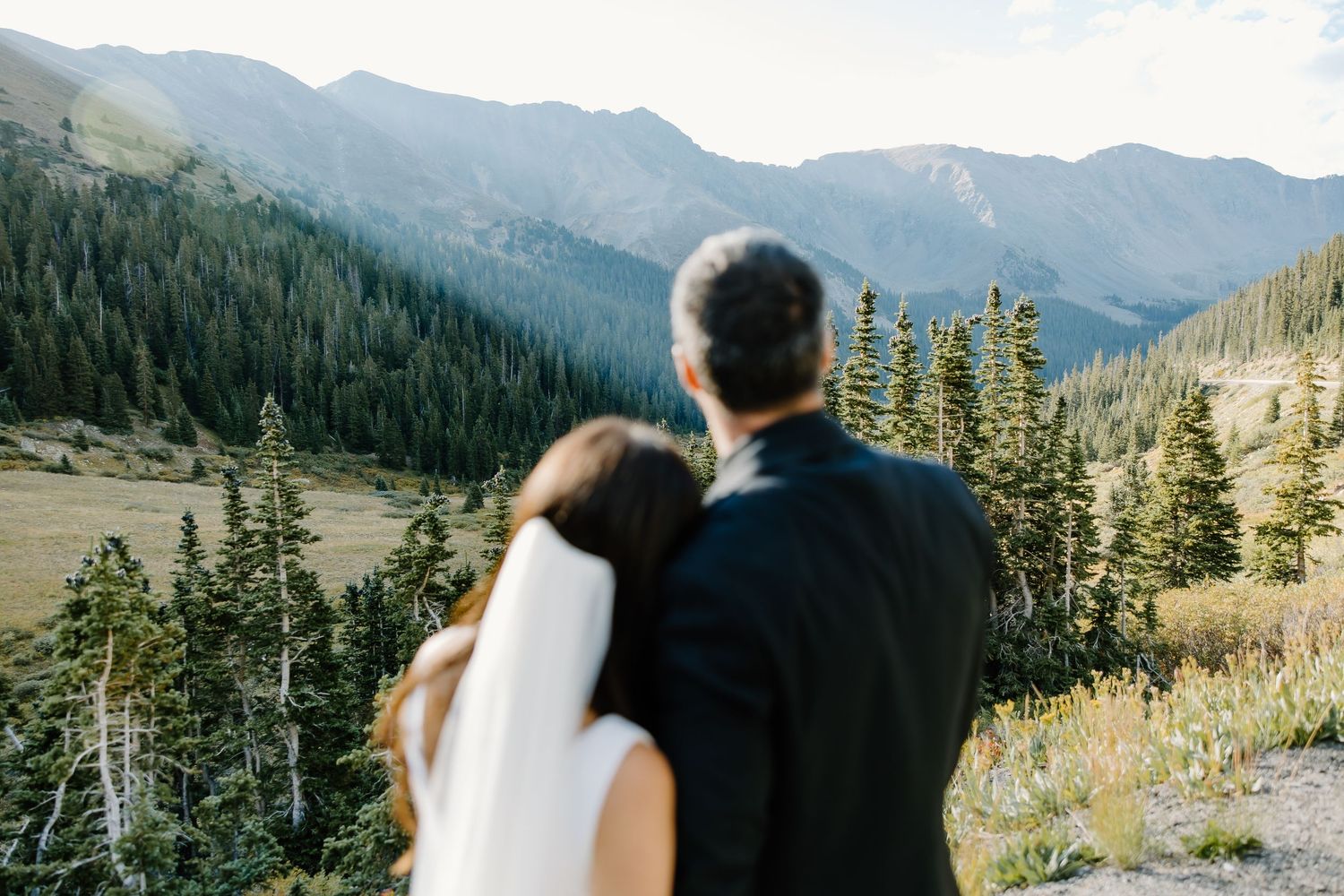 Couple embracing on mountainside with dramatic peaks and forest in background.