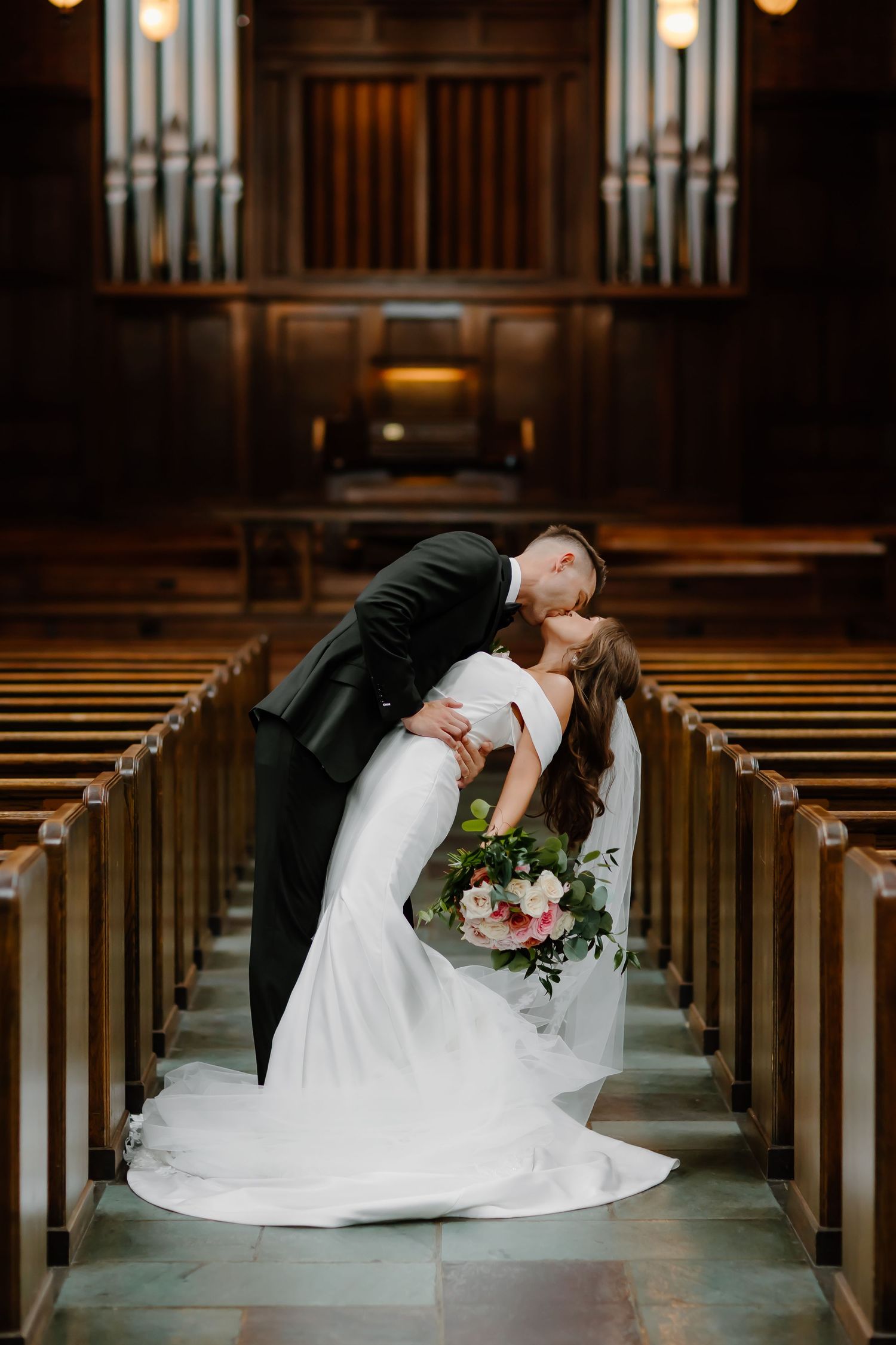 A romantic dip pose captured in a church aisle with wooden pews and organ pipes in the background.