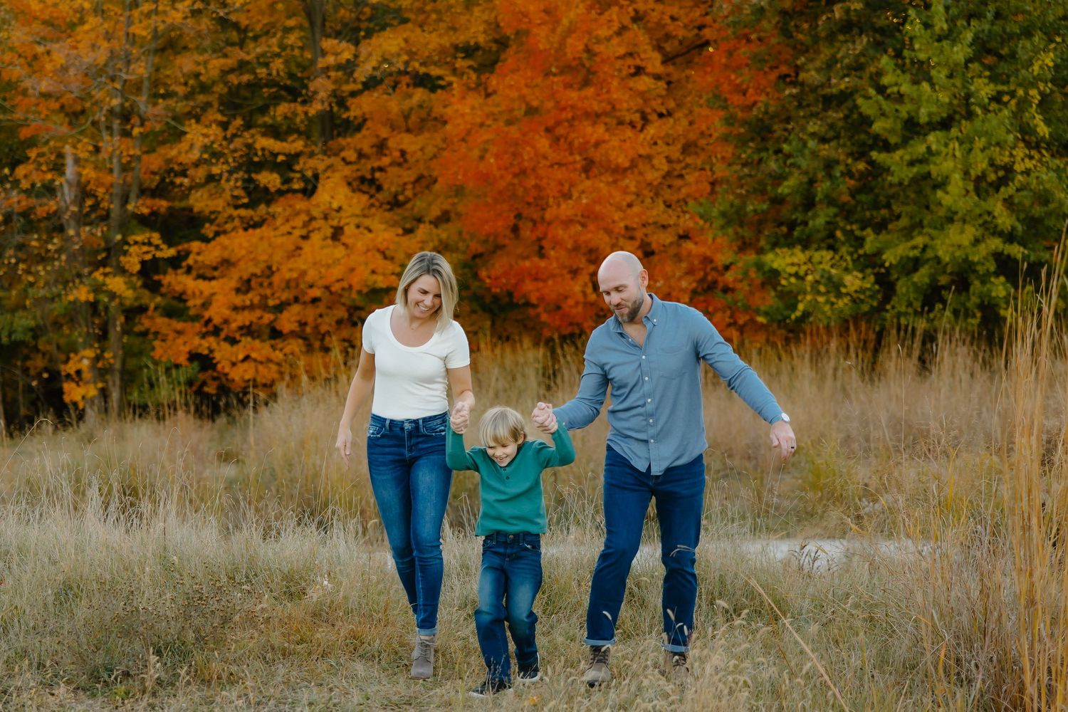 Family members hold hands while walking through tall grass with fall foliage behind them.