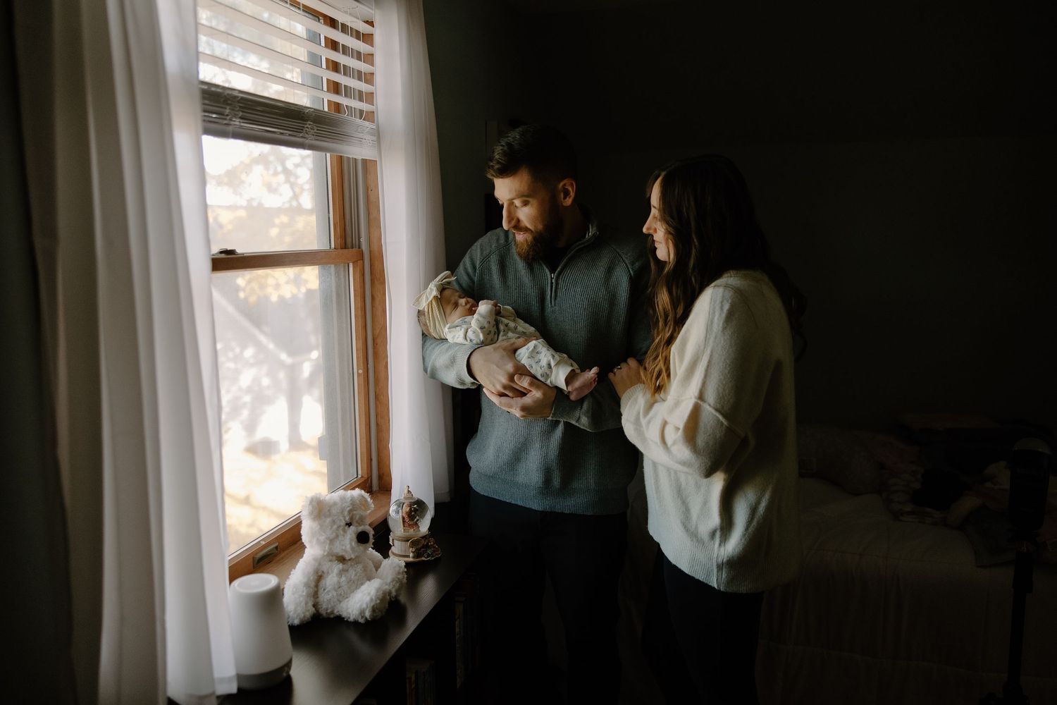 A family stands by a window holding their newborn while their dog looks on.