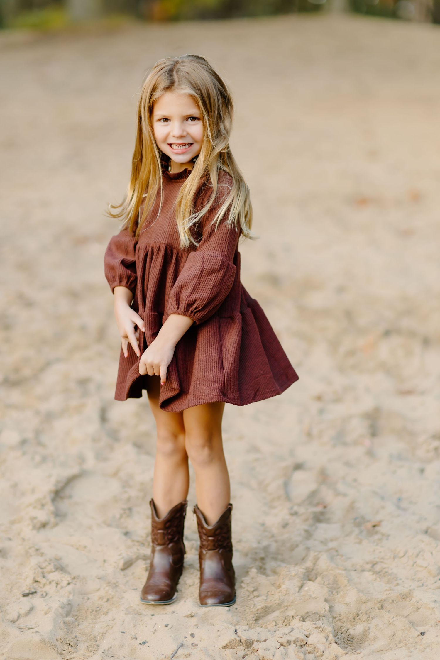 Child in burgundy dress and boots crouches playfully on sandy ground.