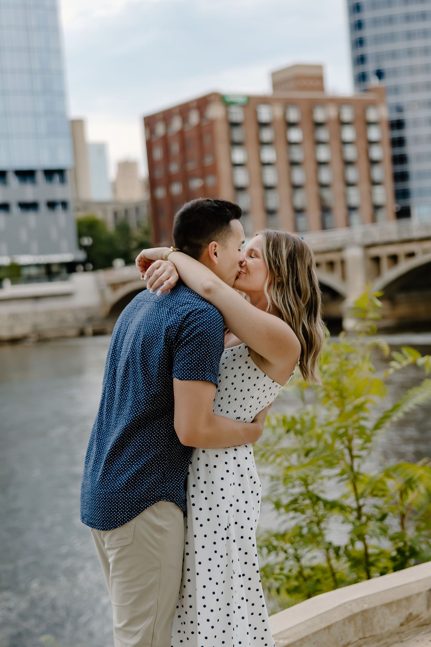 Romantic couple embracing by city river with urban architecture in background.