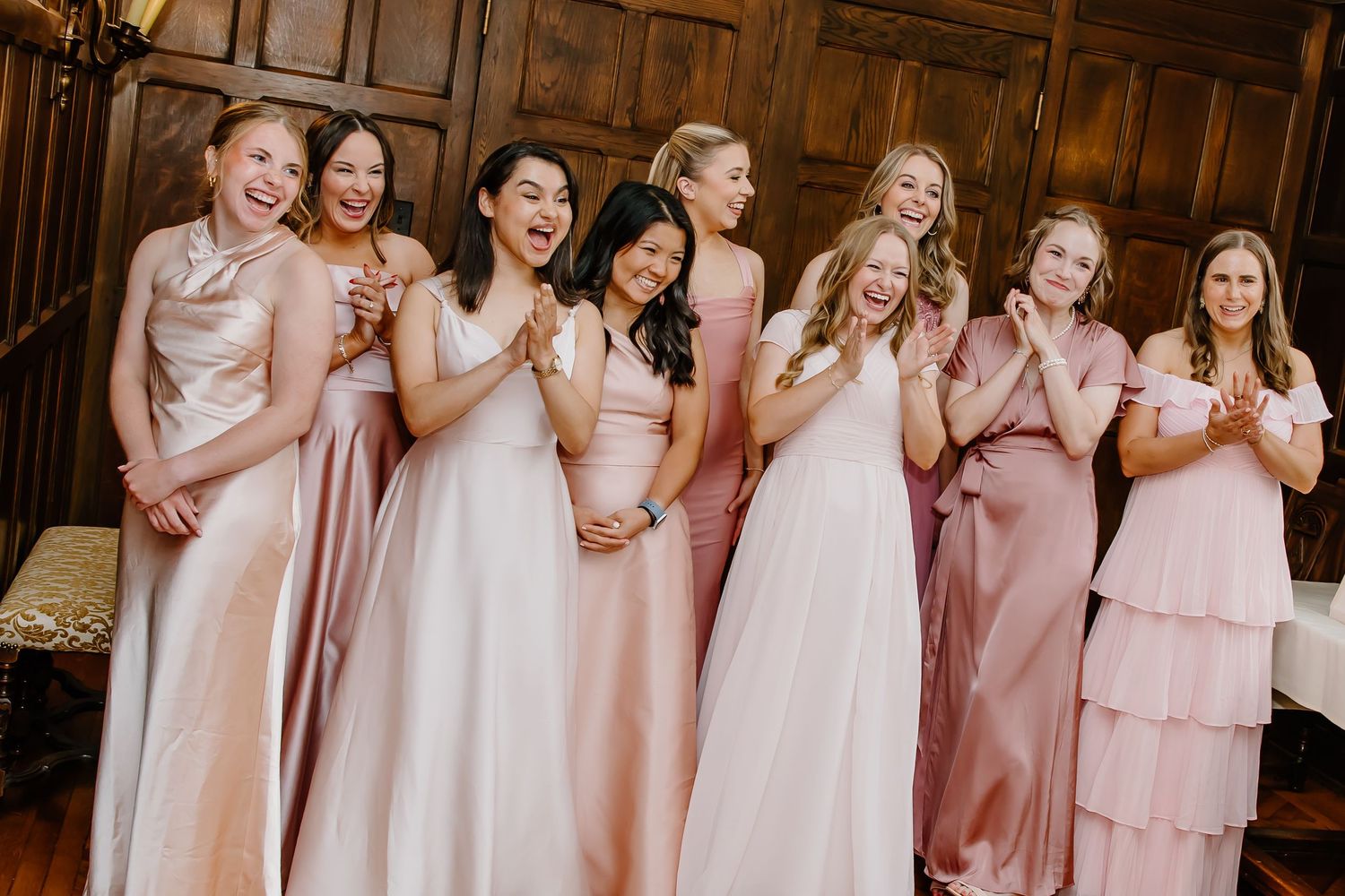 Joyful bridesmaids wearing blush pink dresses share a candid moment in an elegant wood-paneled room.