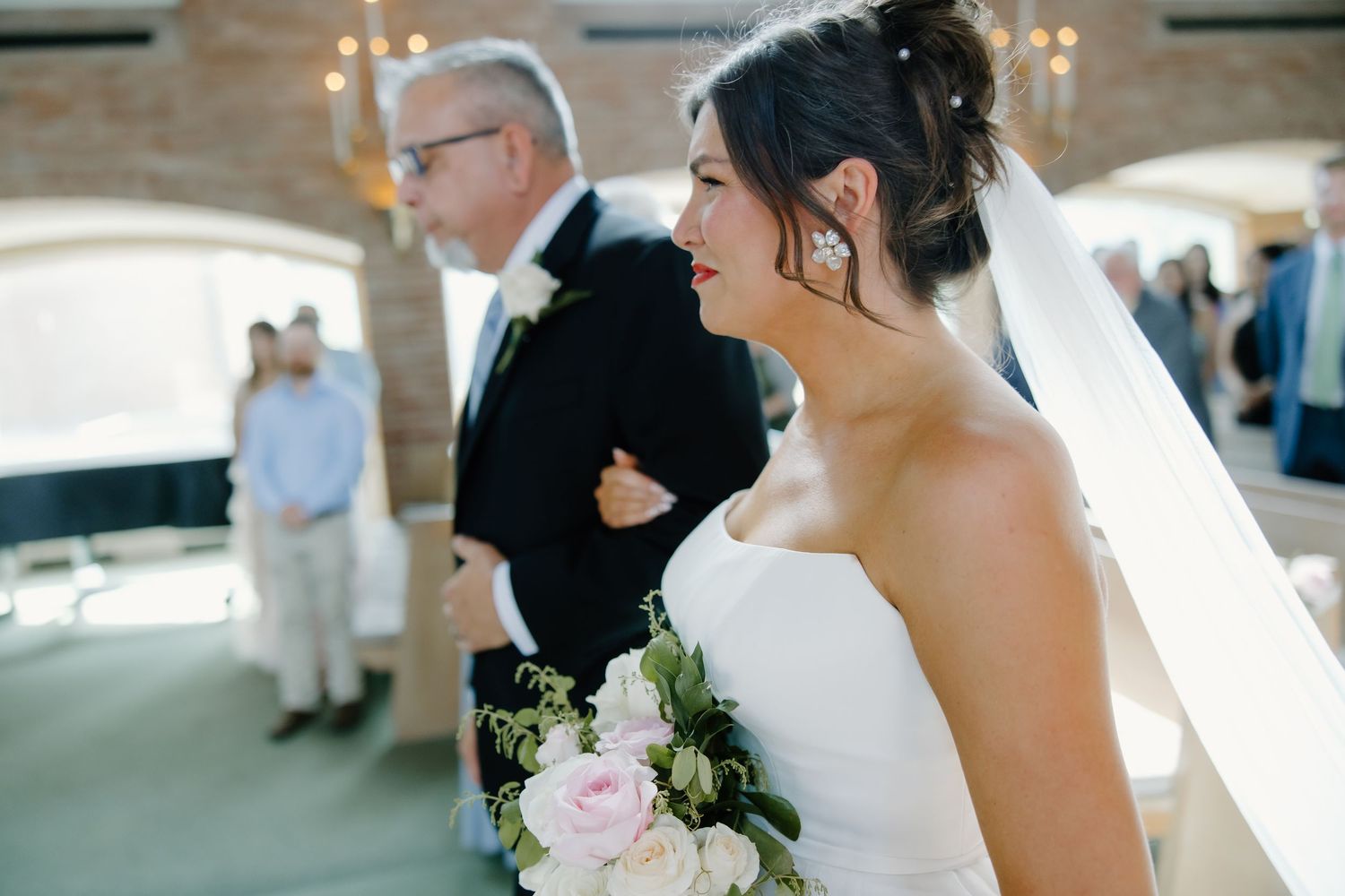 A bride holding a pink and white bouquet smiles during an outdoor wedding ceremony.