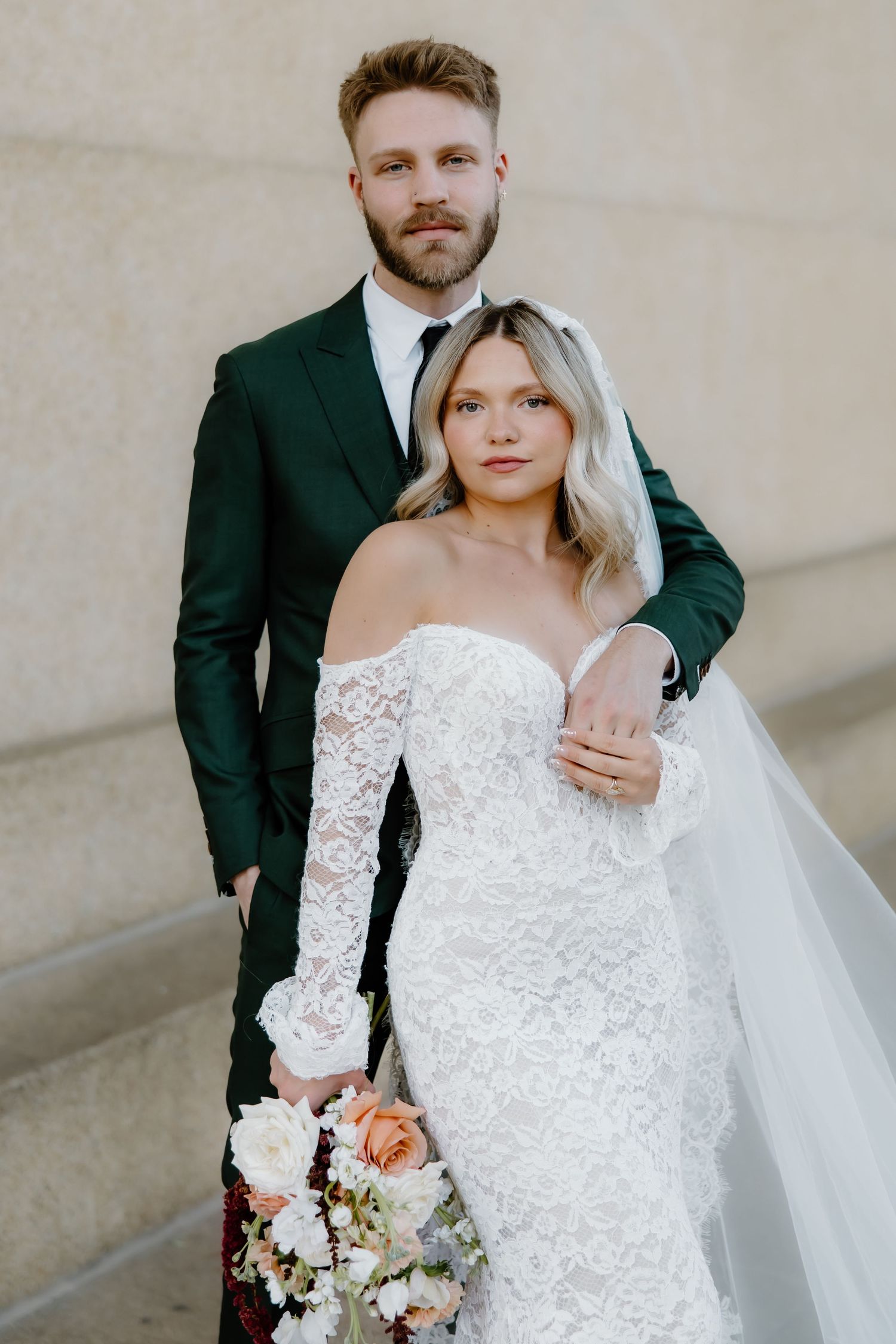 Bride in off-shoulder lace gown holds bouquet beside groom in dark green suit, posing together outdoors.