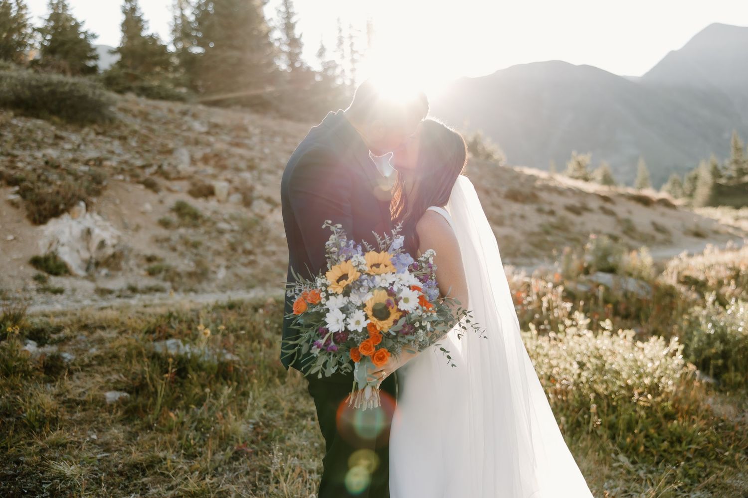 A bride holding a vibrant sunflower bouquet poses against a mountain backdrop at golden hour.