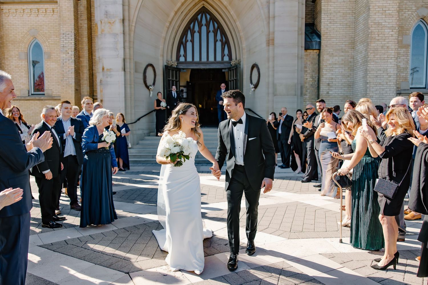 Wedding party and guests gathered outside a historic stone church in bright sunlight.
