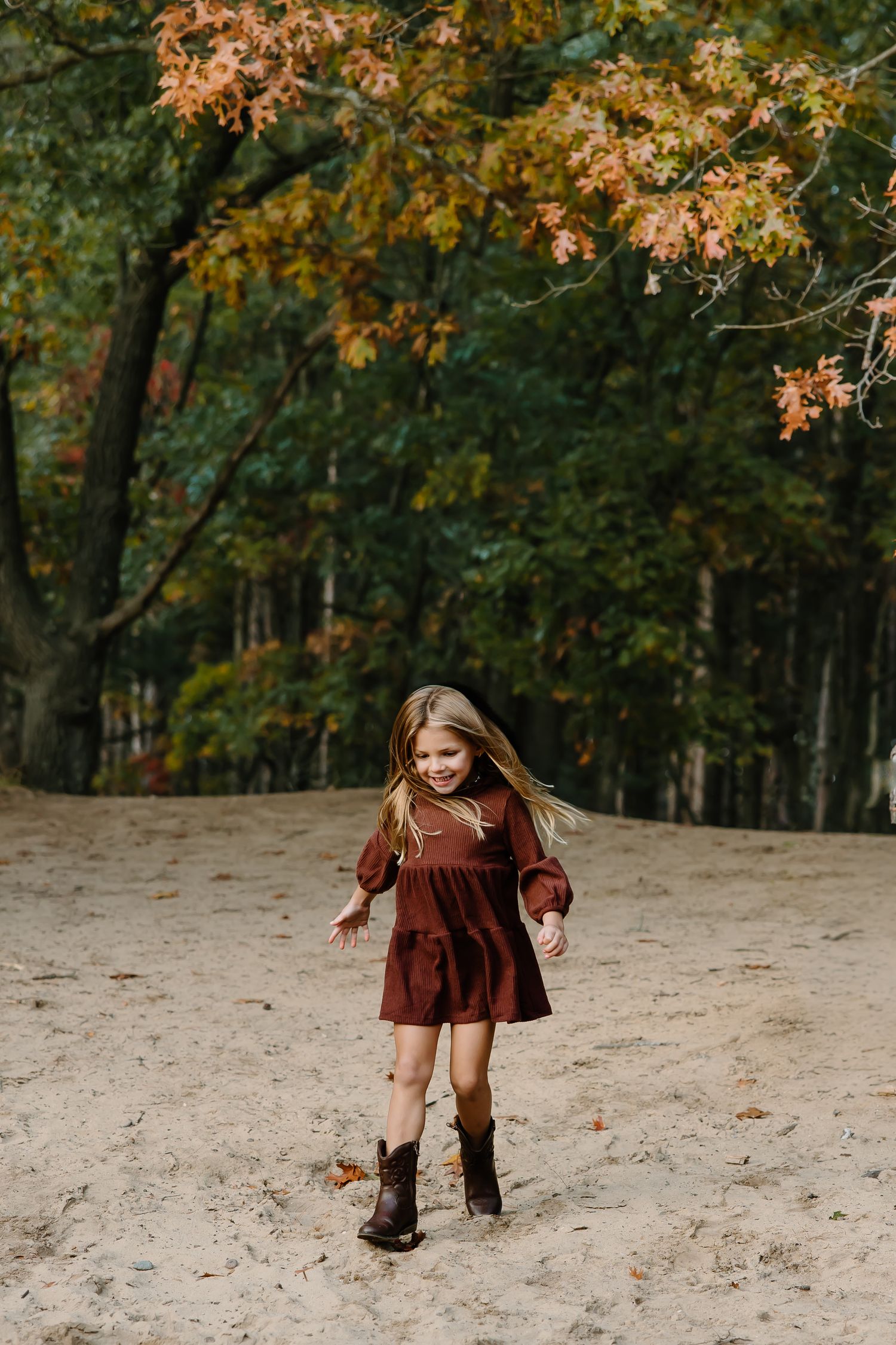Child in burgundy dress plays on sandy slope surrounded by fall foliage.