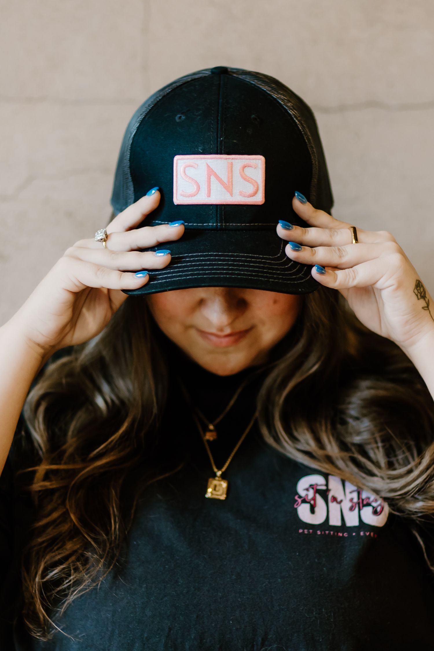 Person wearing a black SNS branded baseball cap and necklace looking down while adjusting their hat.