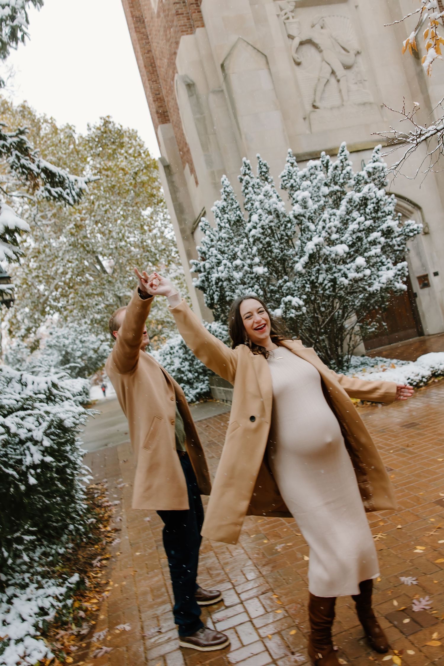 Playful winter maternity photos with snow-covered trees and brick walkway.