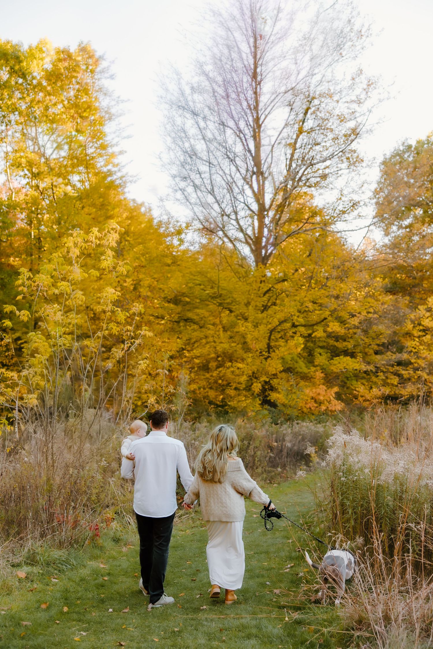 Couple walking on grass path surrounded by golden fall foliage and trees.