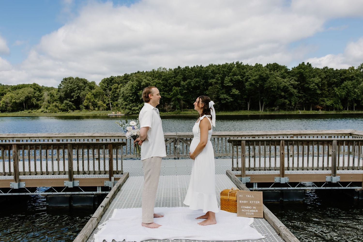 A couple in white outfits stands back-to-back on a lakeside dock during their wedding.