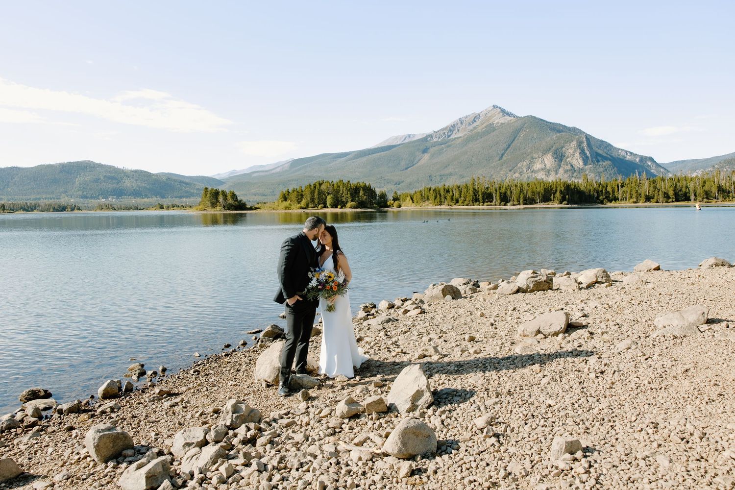 A scenic mountain lake shoreline with rocky beach and evergreen trees under blue skies.