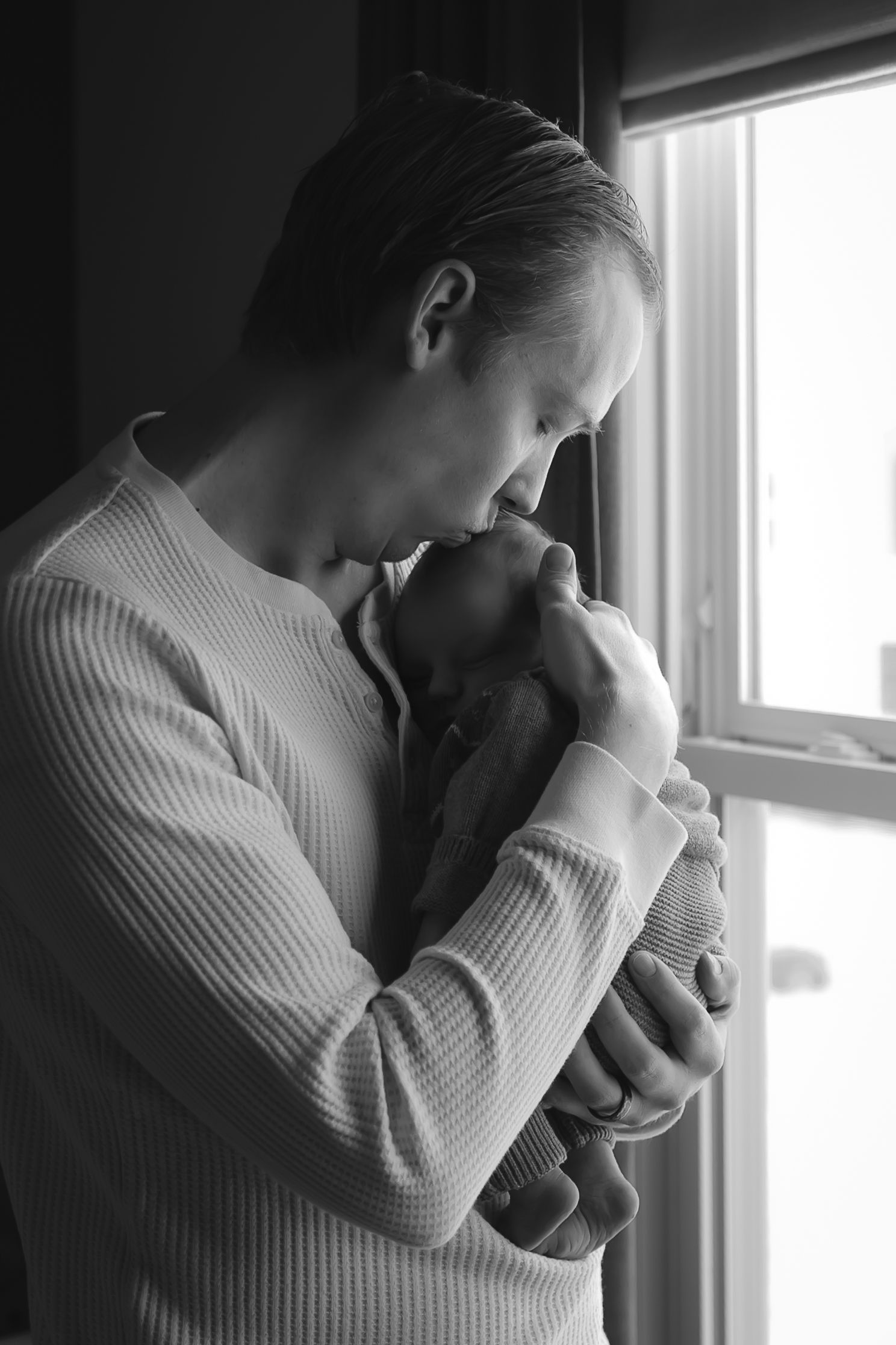 Emotional black and white photos of parent holding newborn by a window.