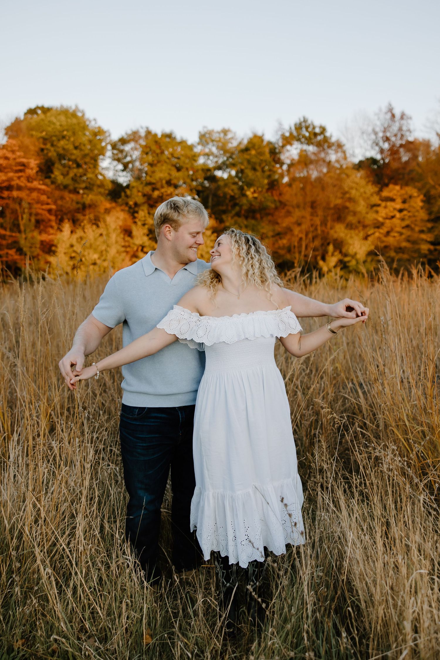 Couple walking through tall autumn grass with golden trees in background.