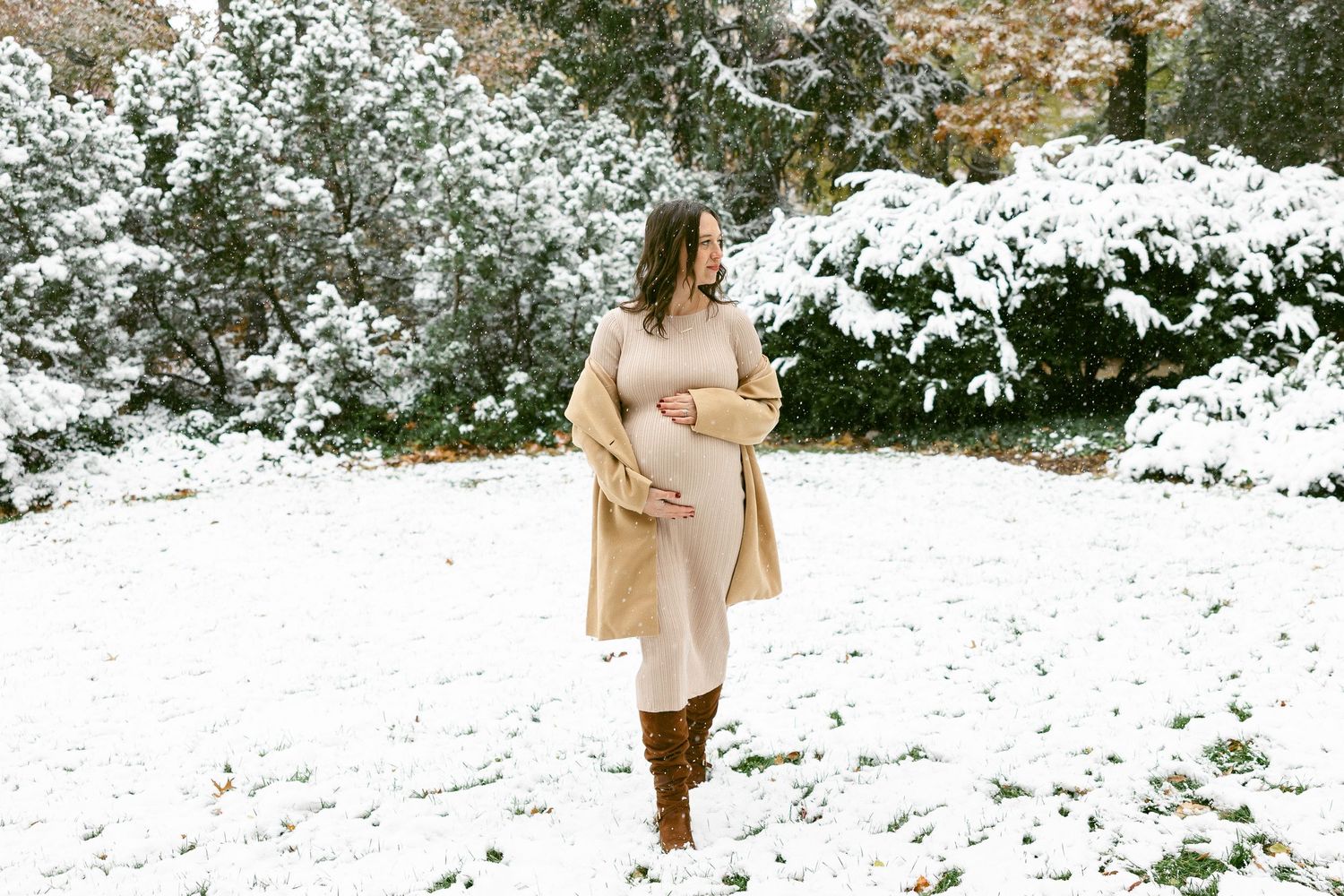 Woman in cream dress and camel coat walking through snow-covered landscape.