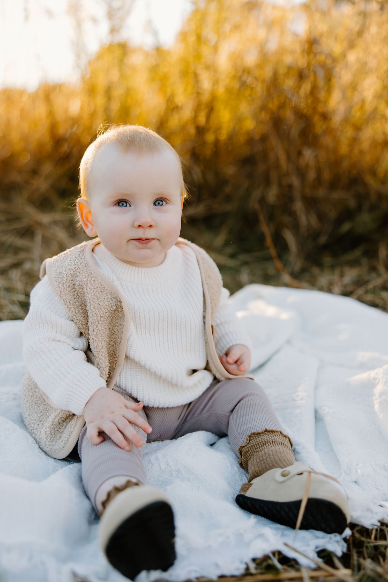A baby in white sweater and beige vest sits on a white blanket outdoors during golden hour.