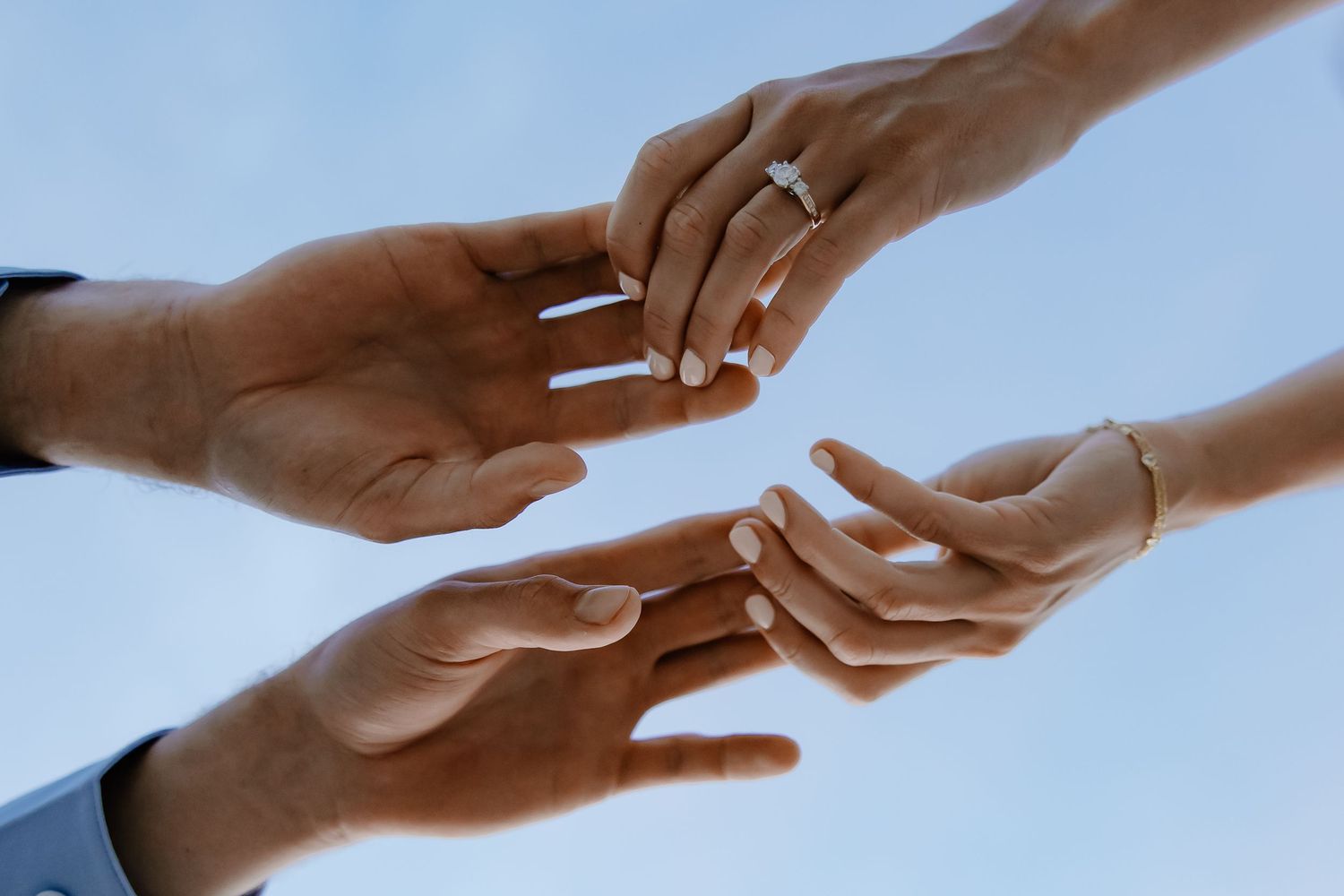 Two hands reaching towards each other wearing engagement rings against a blue sky background.