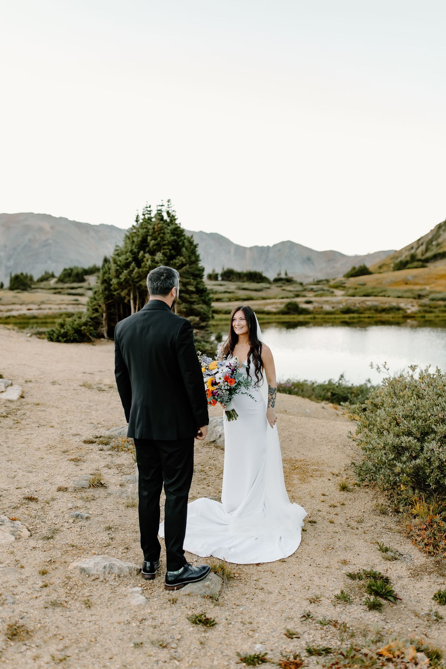 Newlyweds walk together on mountain trail with scenic mountain backdrop.