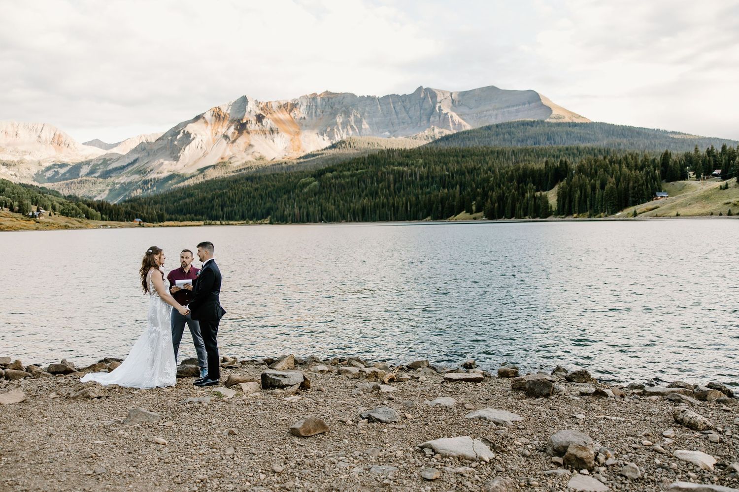 Wedding ceremony unfolds on rocky shoreline with dramatic mountain peaks and lake views.