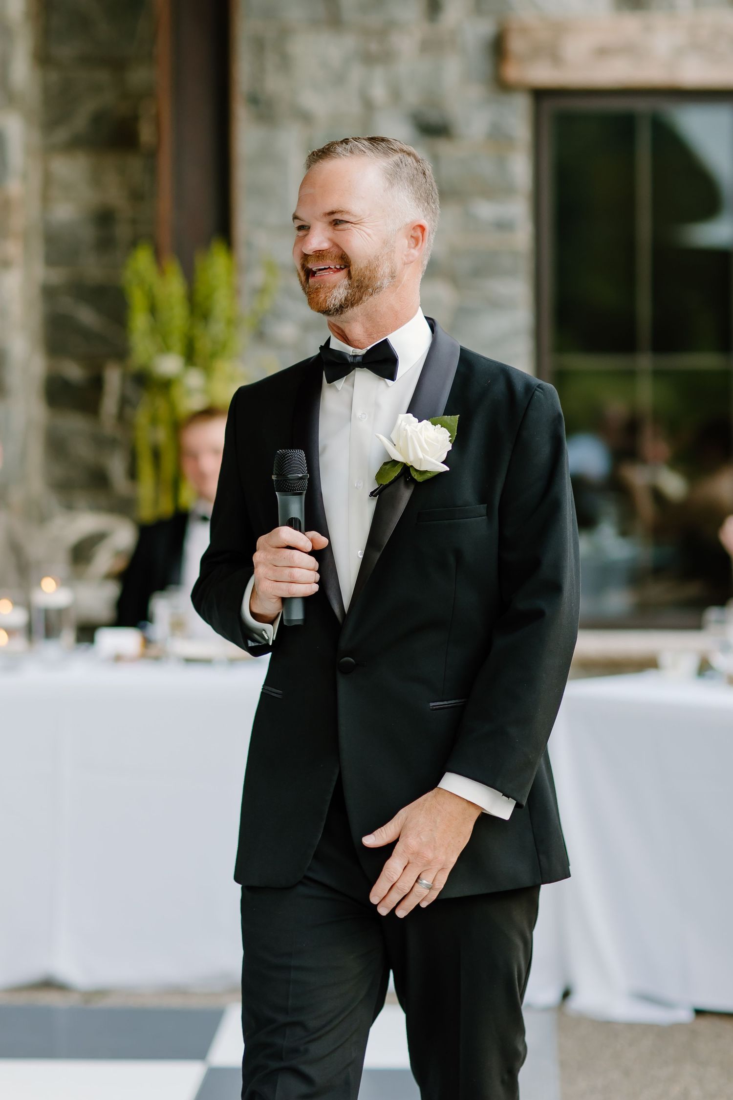 Groom in black tuxedo giving speech during wedding reception.