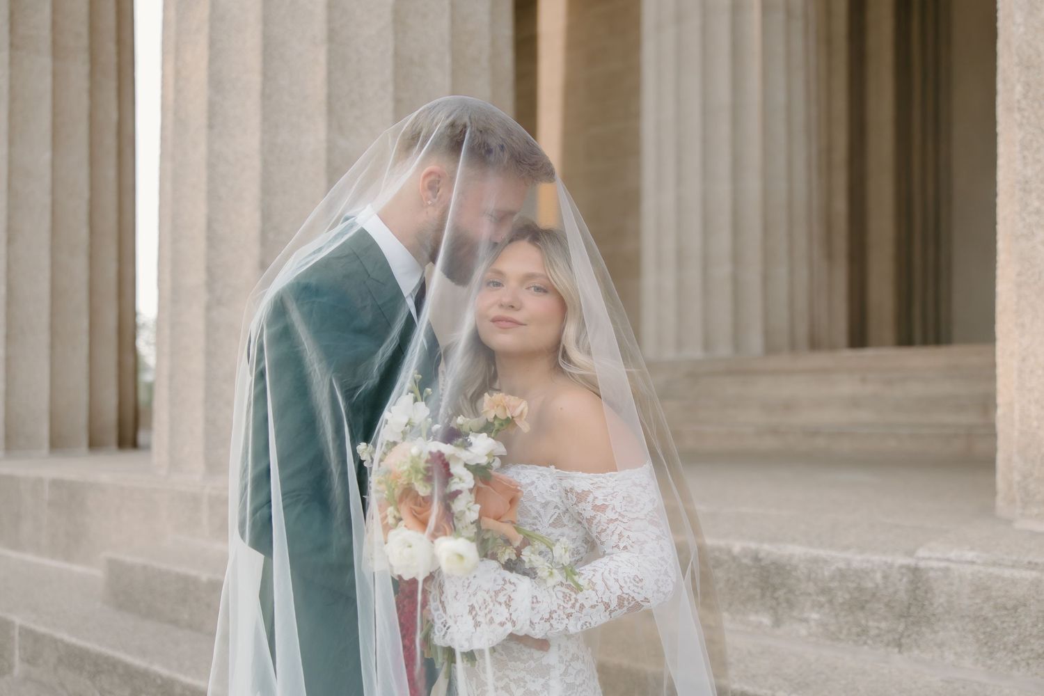 Bride and groom share romantic moment under wedding veil beside elegant stone columns.