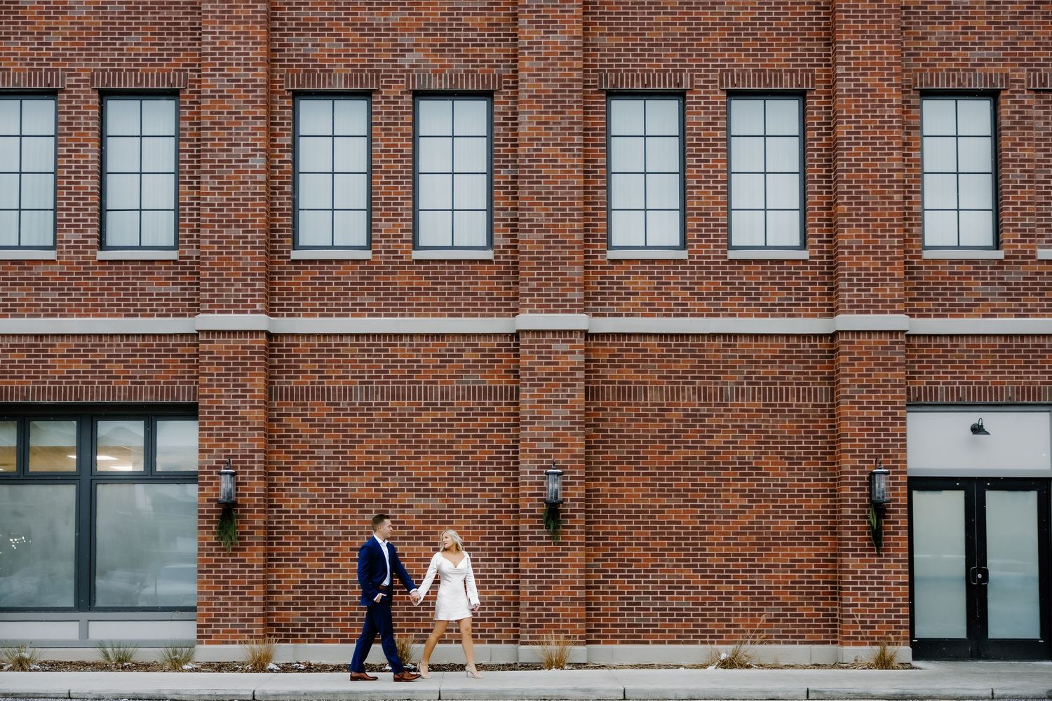 A couple walks along a red brick building exterior with large windows.