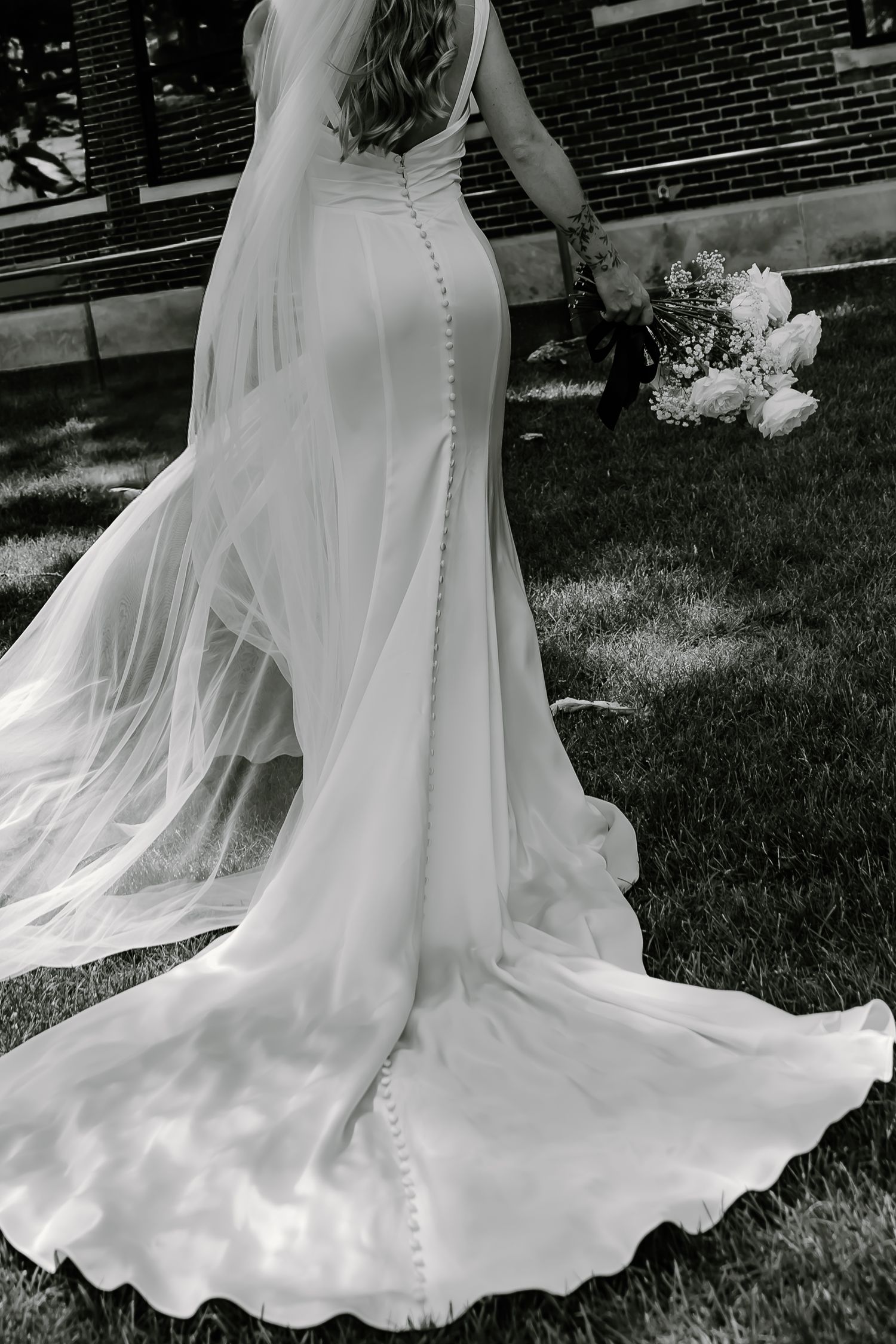 Black and white photograph of a flowing wedding dress train with button details on grass.