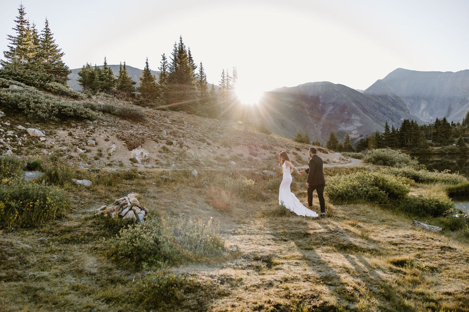 Wedding couple stands on mountain trail at sunset with golden light streaming through evergreens.