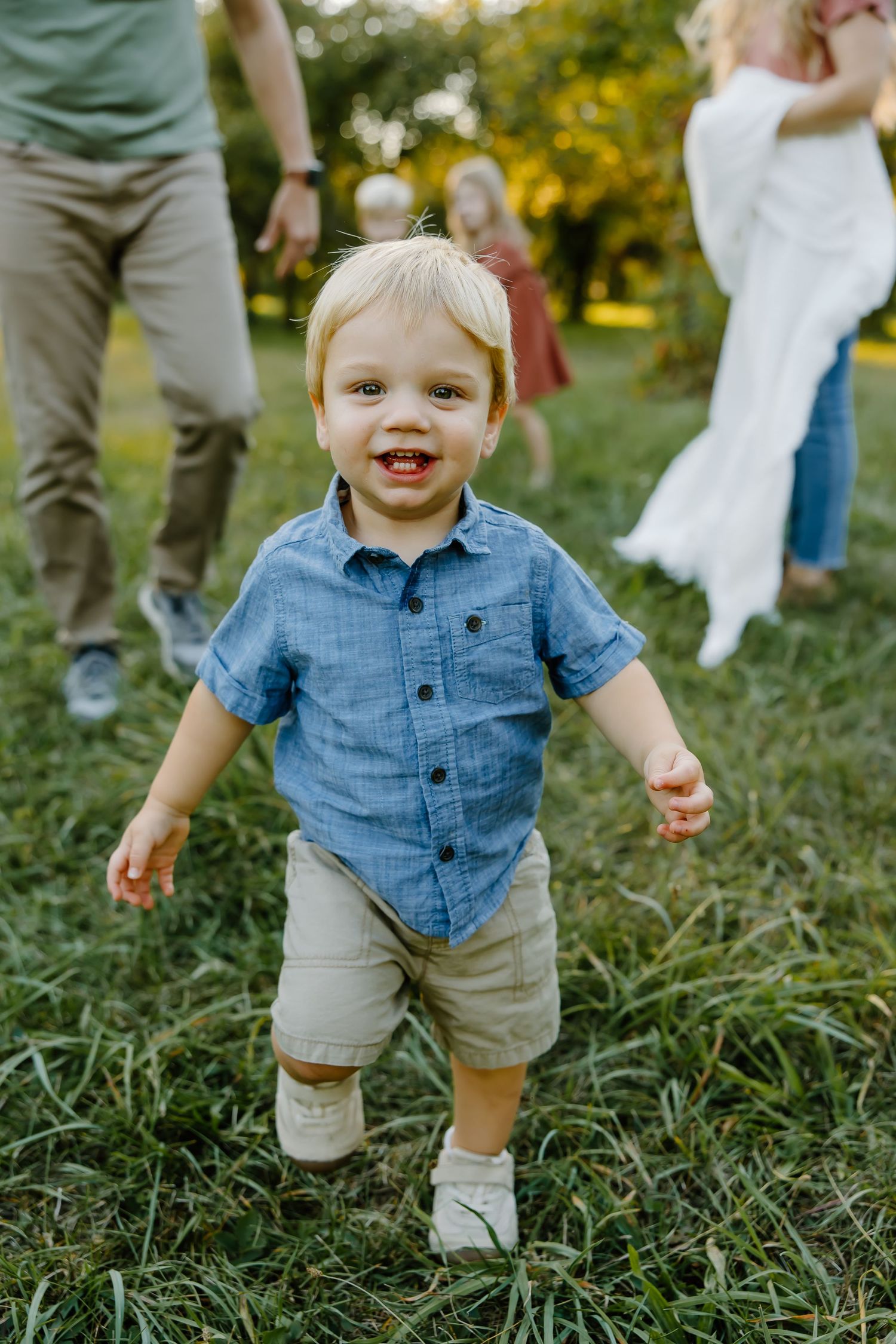 A toddler in denim shirt and khaki shorts smiles while playing outdoors.