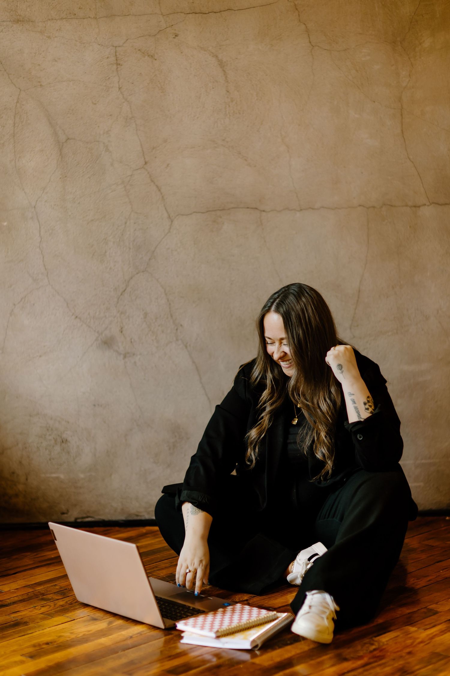 Person in black clothing sits cross-legged on wooden floor working on laptop with notebook nearby in moody setting.