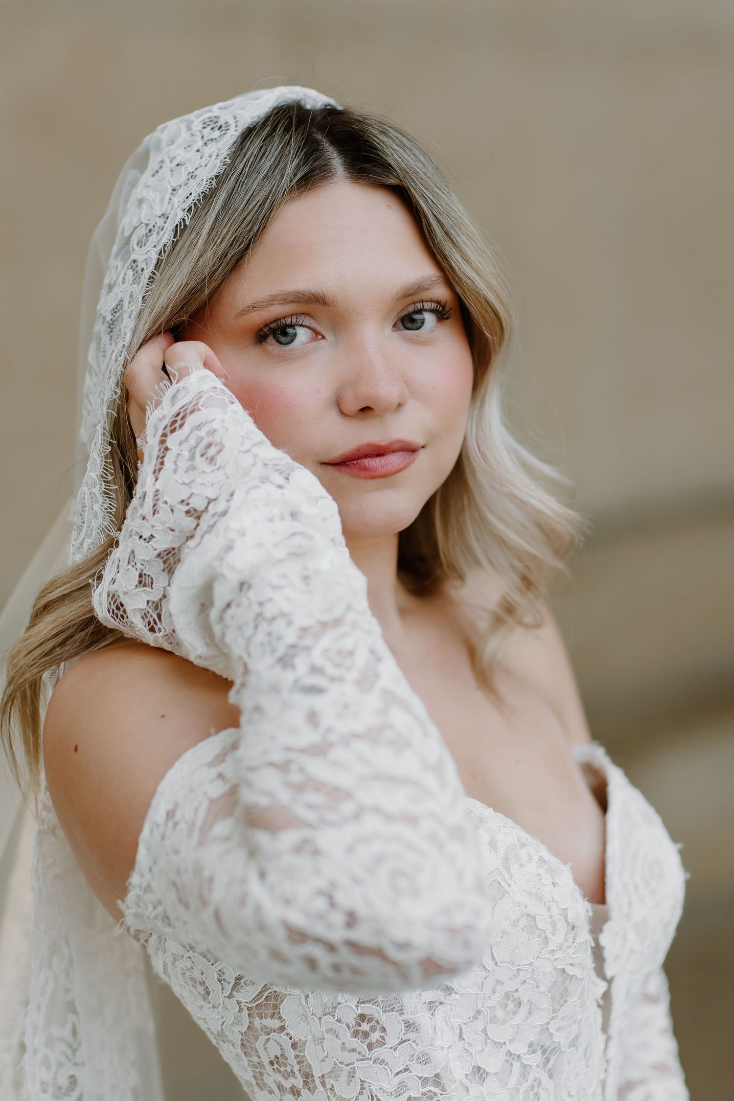 Bride in lace long-sleeve wedding dress and veil, posing elegantly with hand near face, soft natural light.