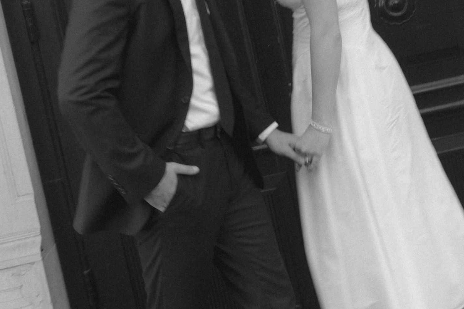 Black and white photo of a bride and groom holding hands in elegant wedding attire outside a door.