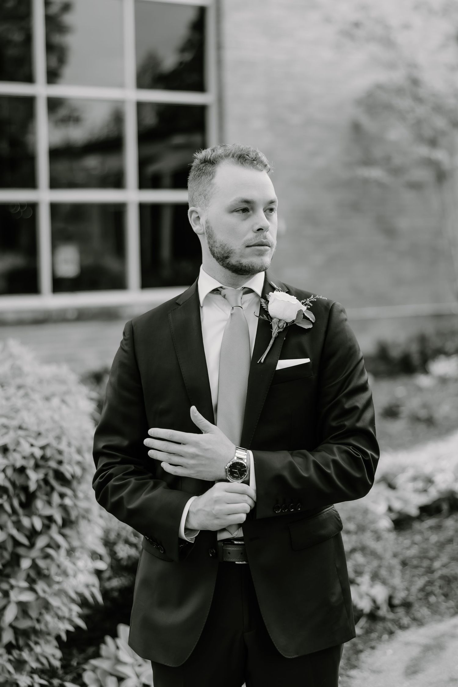 Groom in classic black suit with light tie and boutonniere poses outdoors.