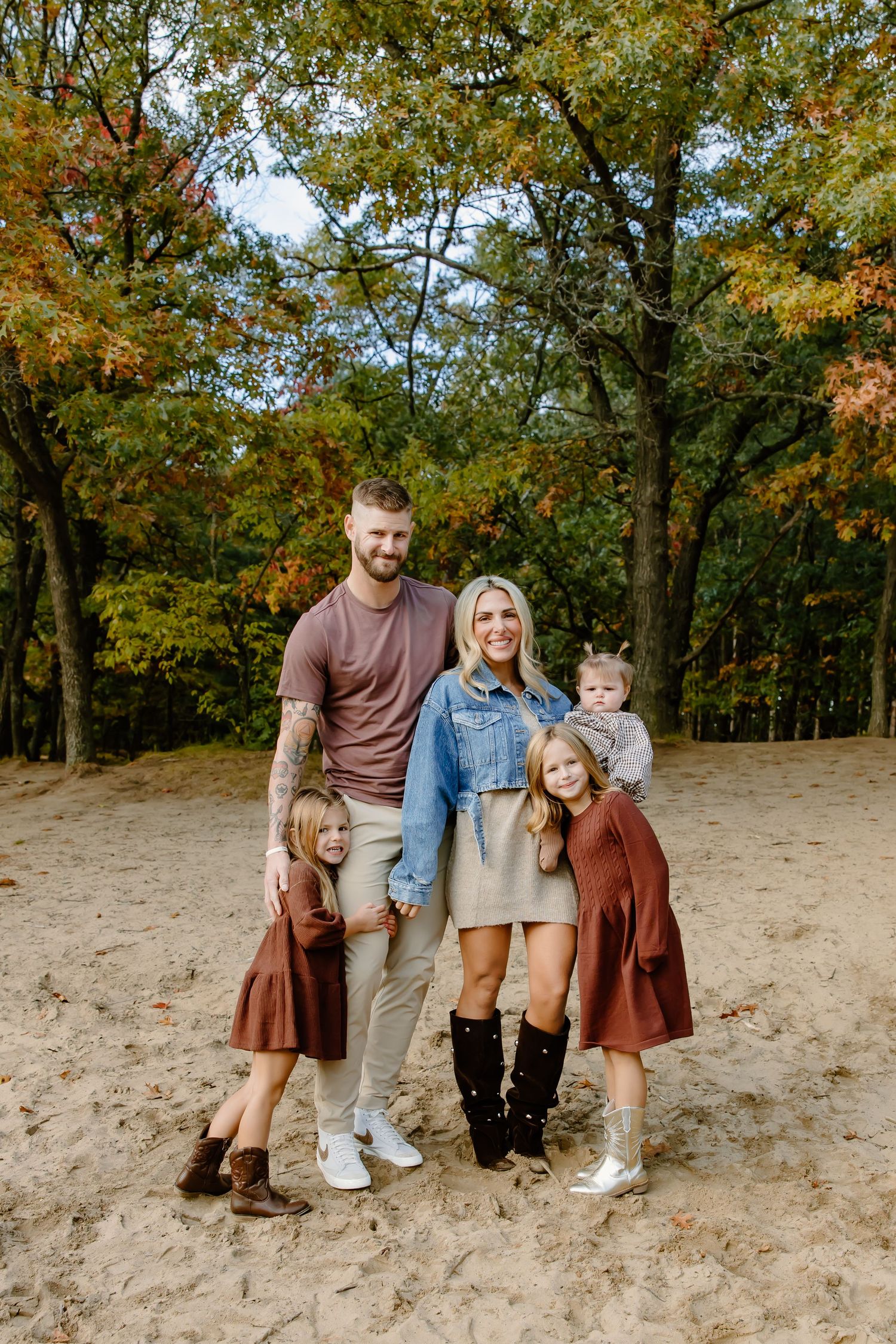 Family group poses on sandy slope with autumn trees in background wearing matching casual attire.