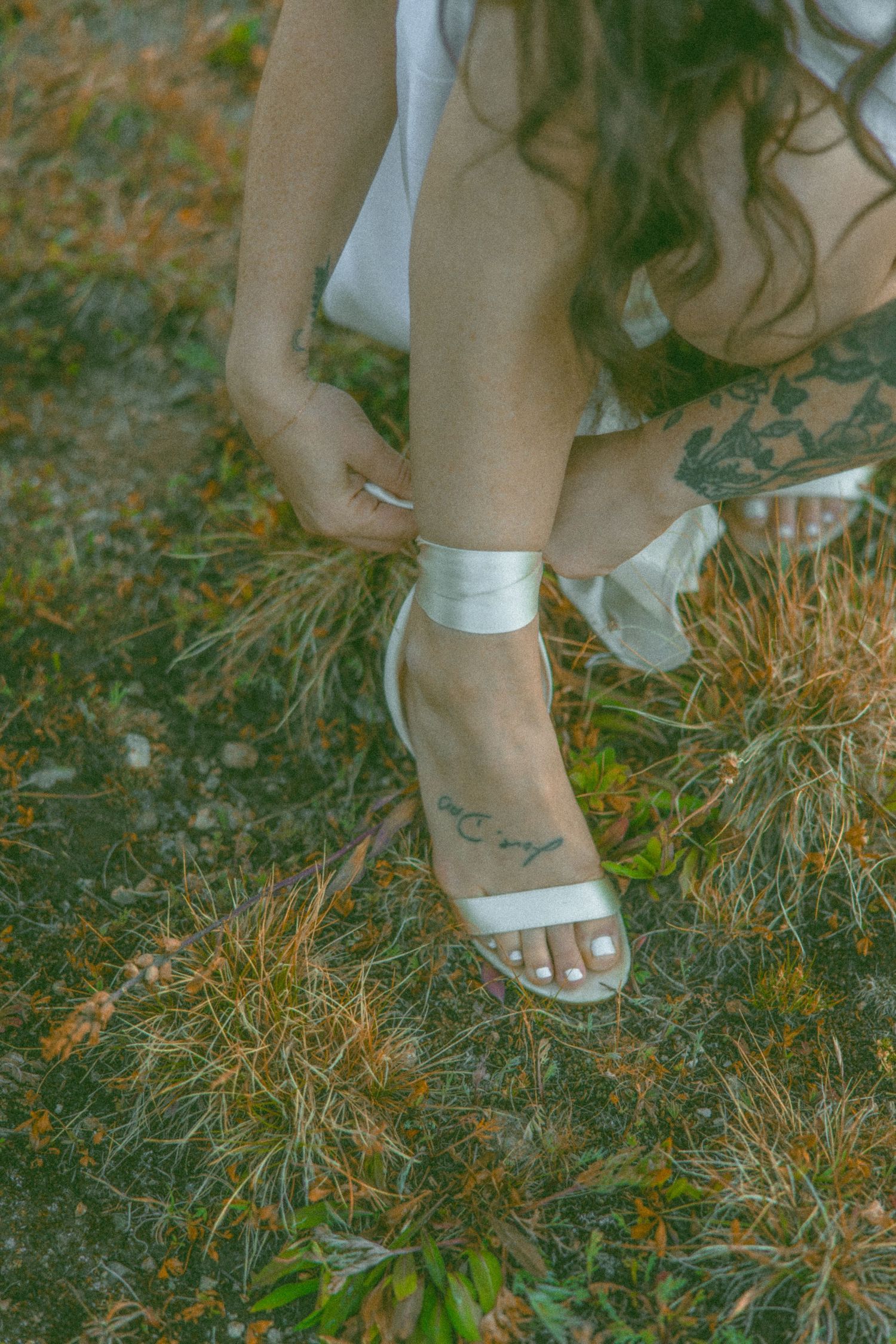 Close-up of white wedding sandal and ankle ribbon on grass with small foot tattoo visible.