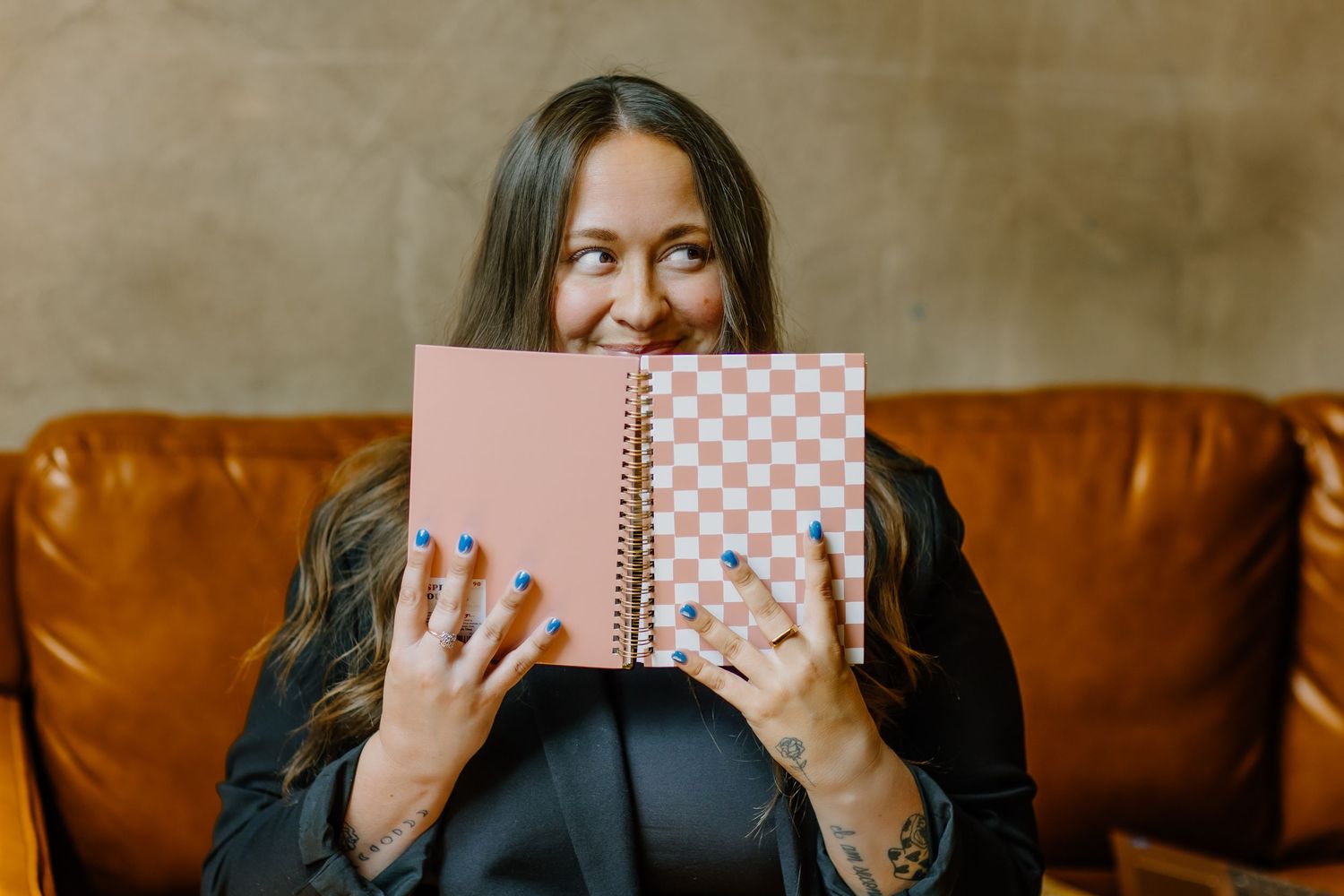 Someone sitting on a brown leather couch holds up a pink spiral notebook to partially cover their face.